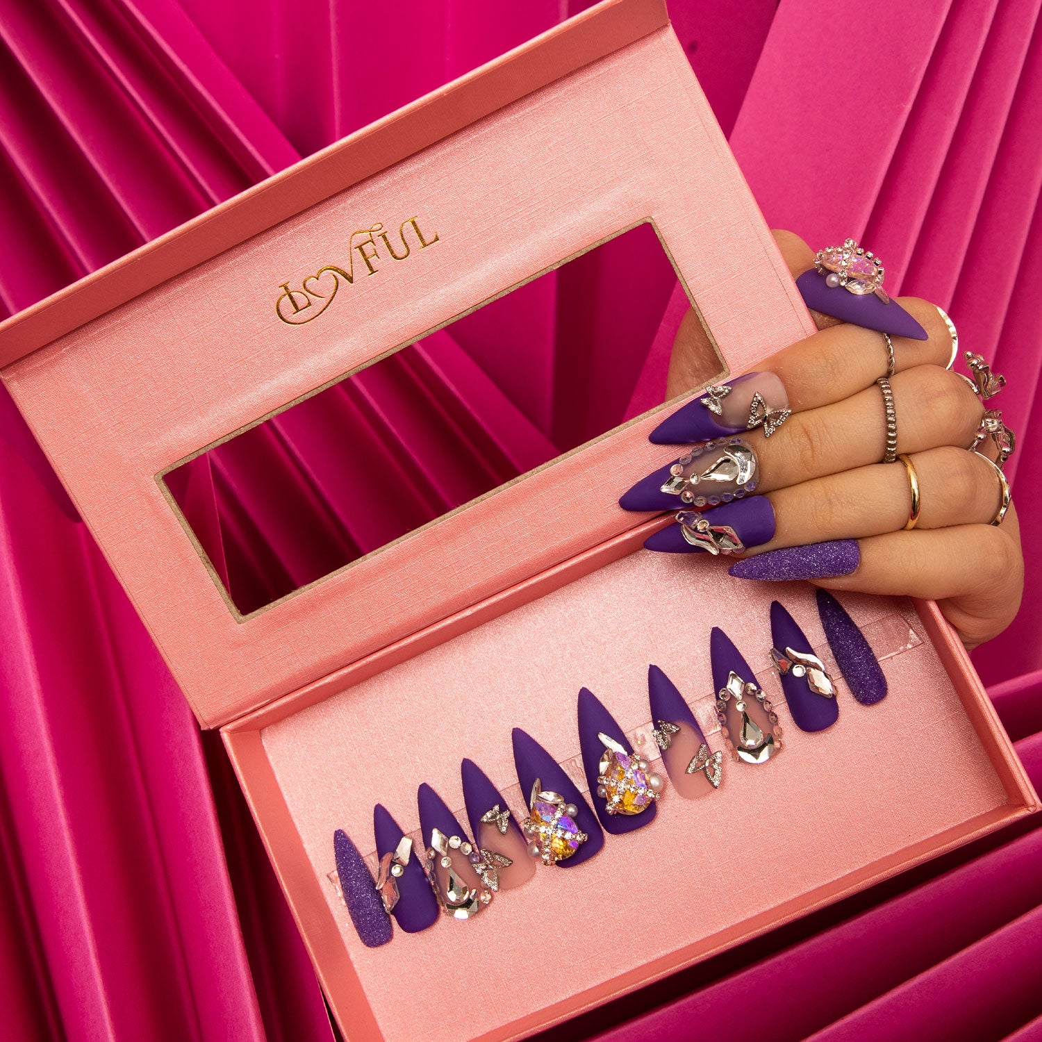 Hand with well-manicured nails holding an open pink box containing a 'Mystique Veil' press-on nail set. The nails are deep purple with butterfly decorations, gem accents, and glitter overlays, set against a pink fabric backdrop.