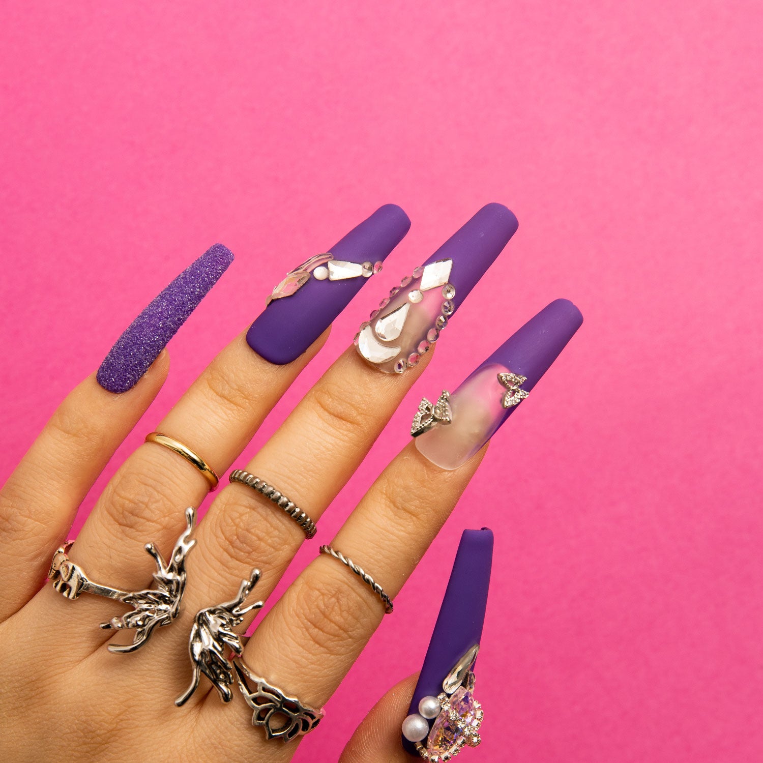 Hand with 'Mystique Veil' deep purple coffin press-on nails adorned with butterfly decorations, gem accents, and glitter overlay, set against a bright pink background.