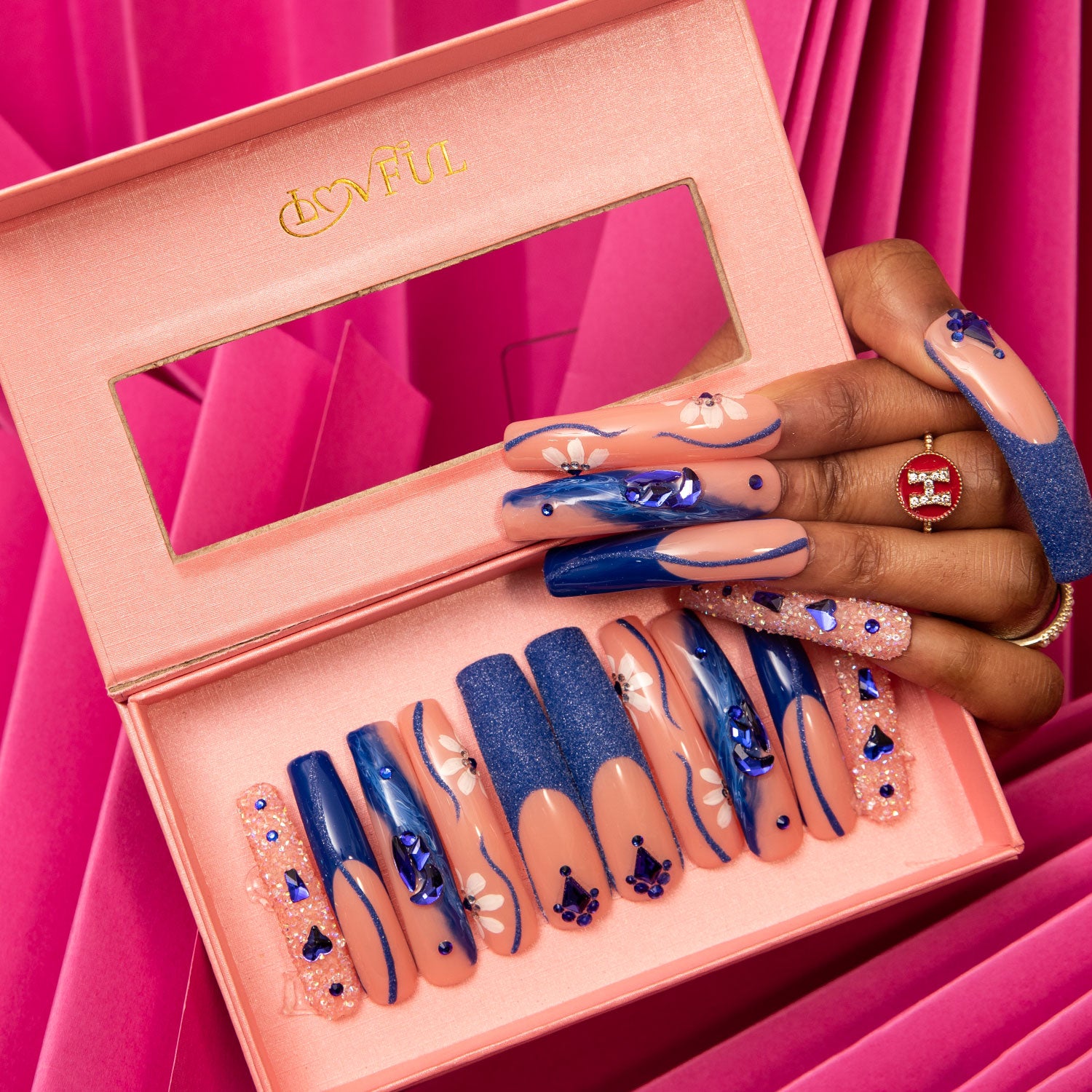 Set of 'Blue Suede' press-on nails in a pink Lovful box with crystal-like decorations and blue heart-shaped gems, held by a hand with matching nails against a pink background.