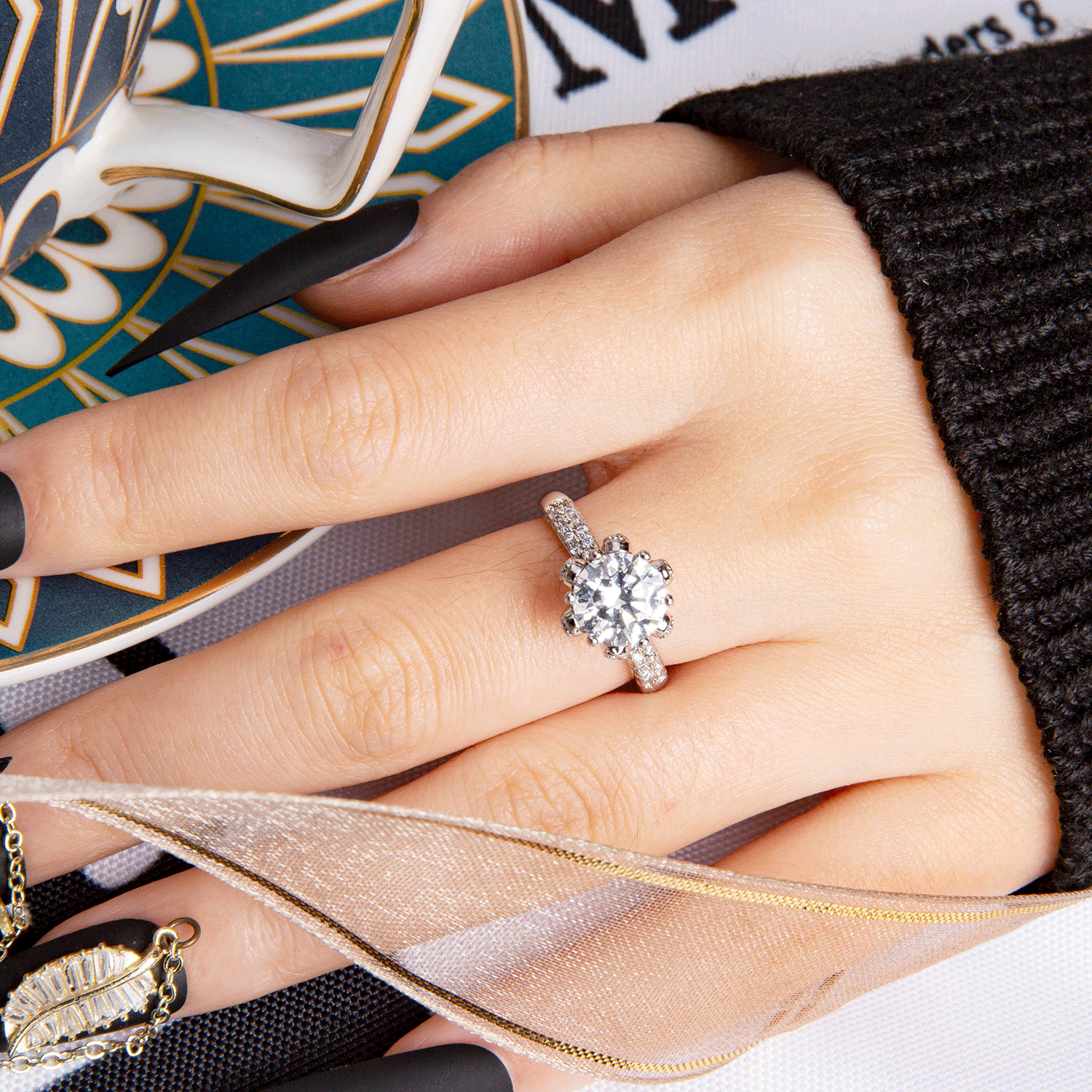 Hand with black matte press-on acrylic nails holding a teacup and wearing a large diamond ring on a silver band. Decorated with a ribbon and a decorative leaf ornament.