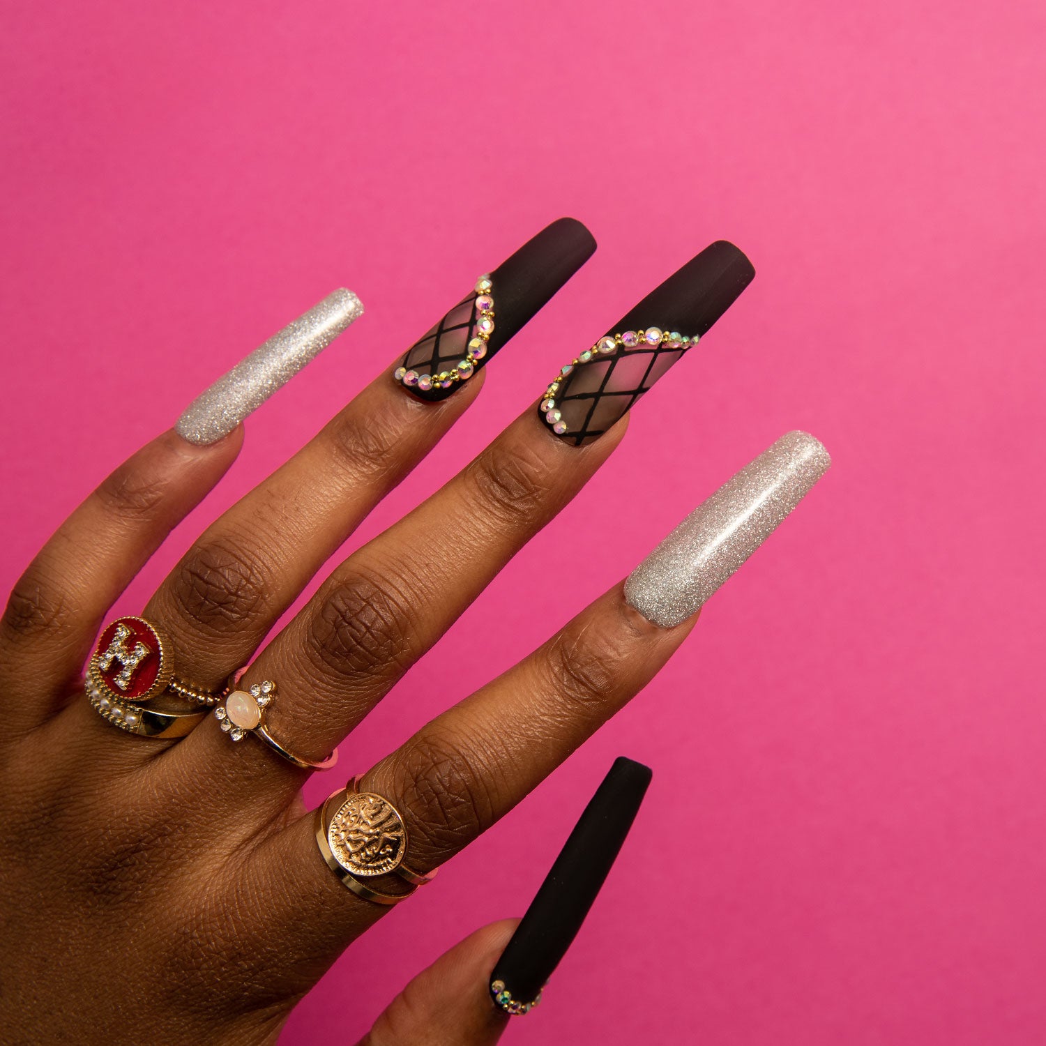 Close-up of a hand with long, square-shaped Black Lace press-on nails. The nails have a black design with silver shimmer and lace decor, accented with small rhinestones. The hand also showcases several rings, set against a pink background.