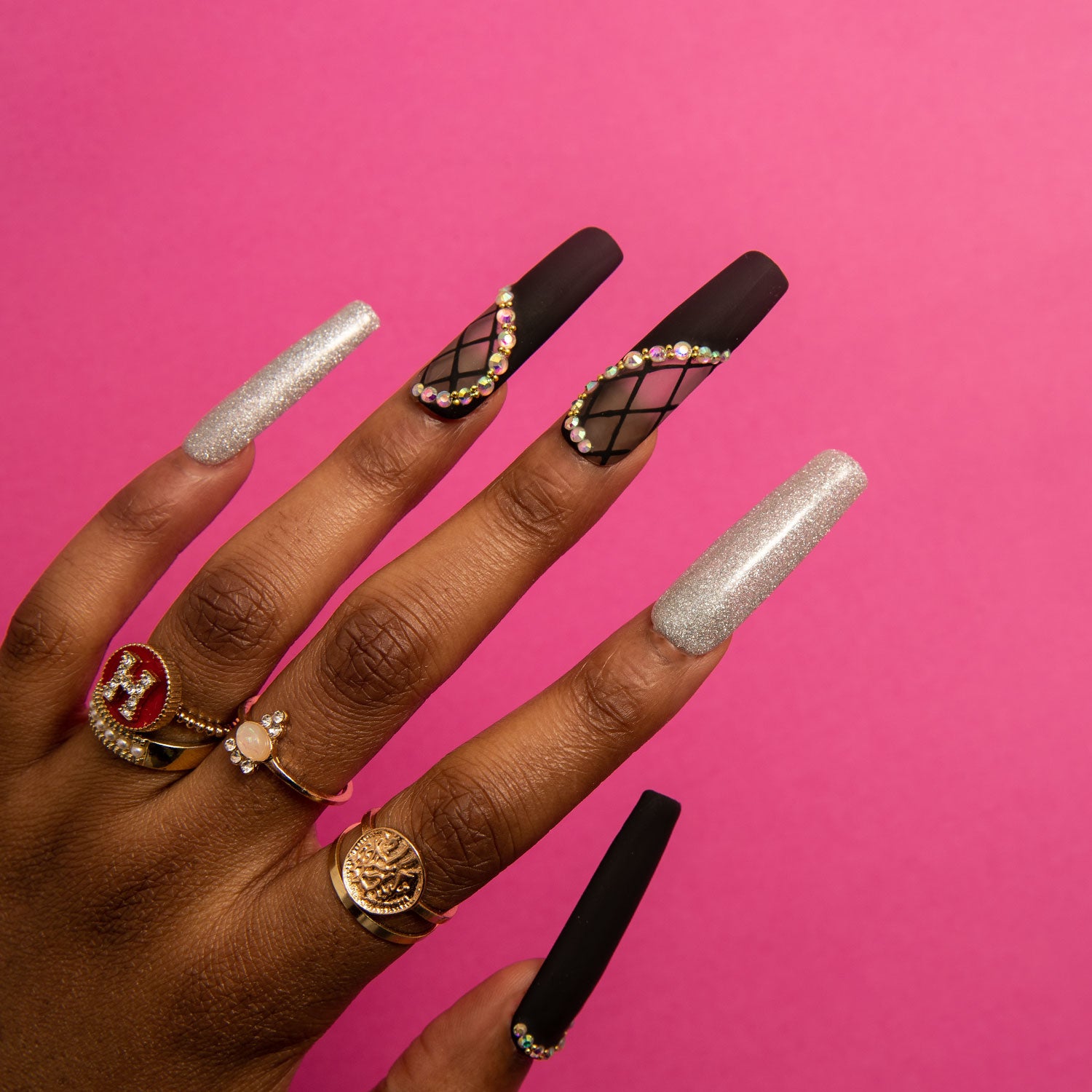 Long square black matte press-on nails with intricate lace designs and rhinestone accents on middle fingers, silver glitter polish on thumb and pinky, worn on hand with gold rings and pink background.