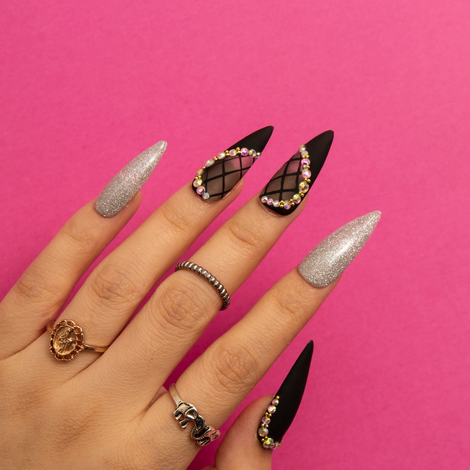 Hand with Black Lace Stiletto press-on nails from Lovful.com. Nails are decorated with silver shimmer, lace patterns, and rhinestone accents, set against a pink background.