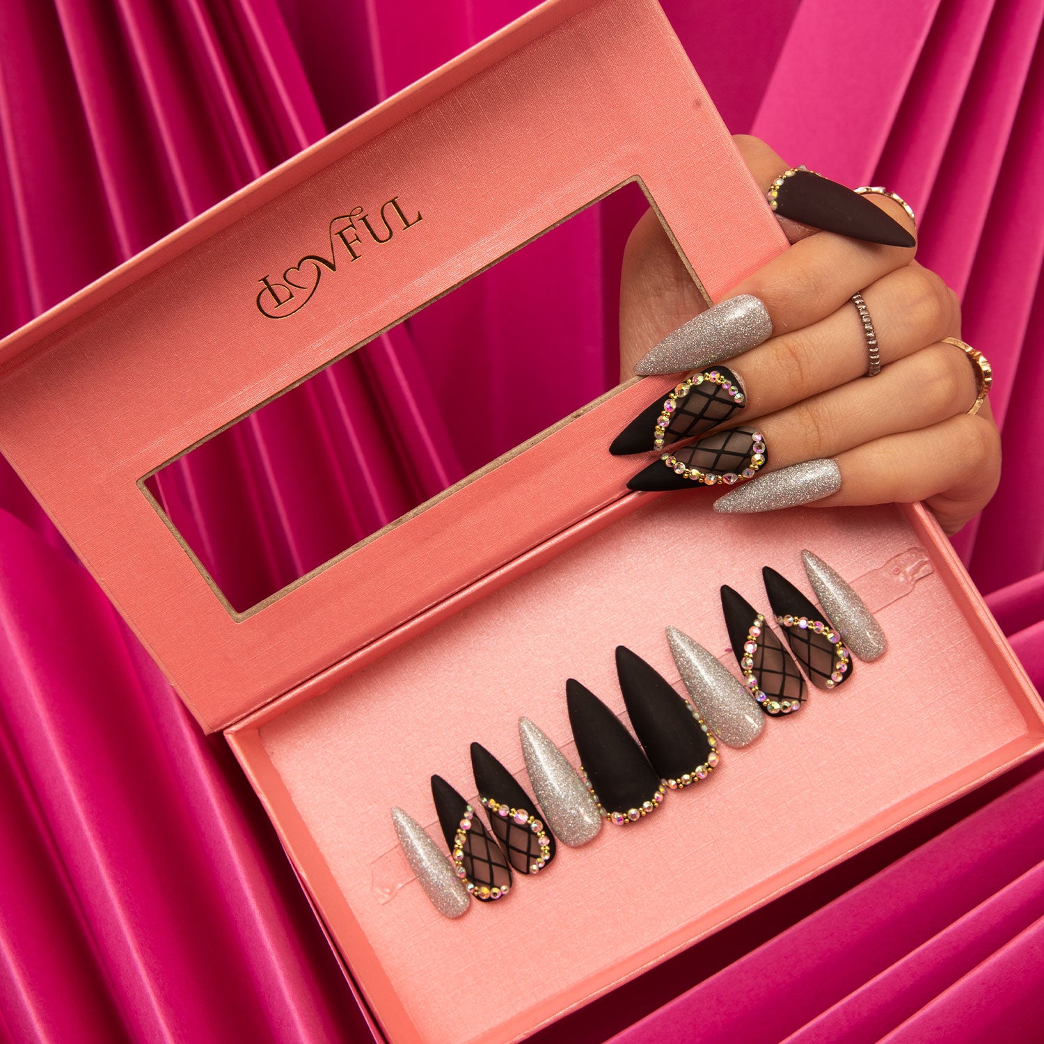 Box of Lovful Black Lace press-on stiletto nails with alternating black and silver shimmer designs, featuring rhinestones and lace decor, held by a hand against a pink fabric background.