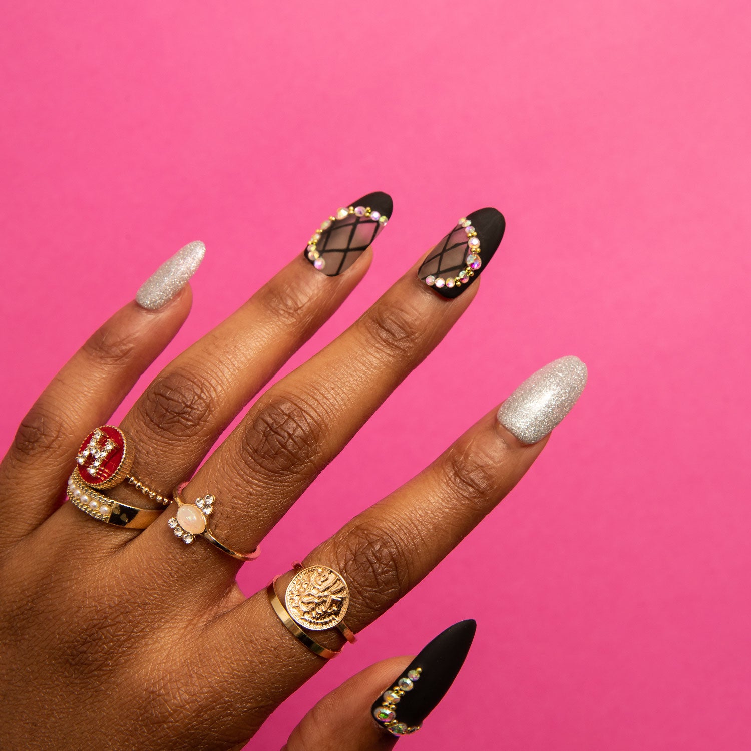 Hand wearing Black Lace press-on nails with black, silver shimmer, lace decor, and rhinestone accents, featuring gold and gemstone rings against a pink background.