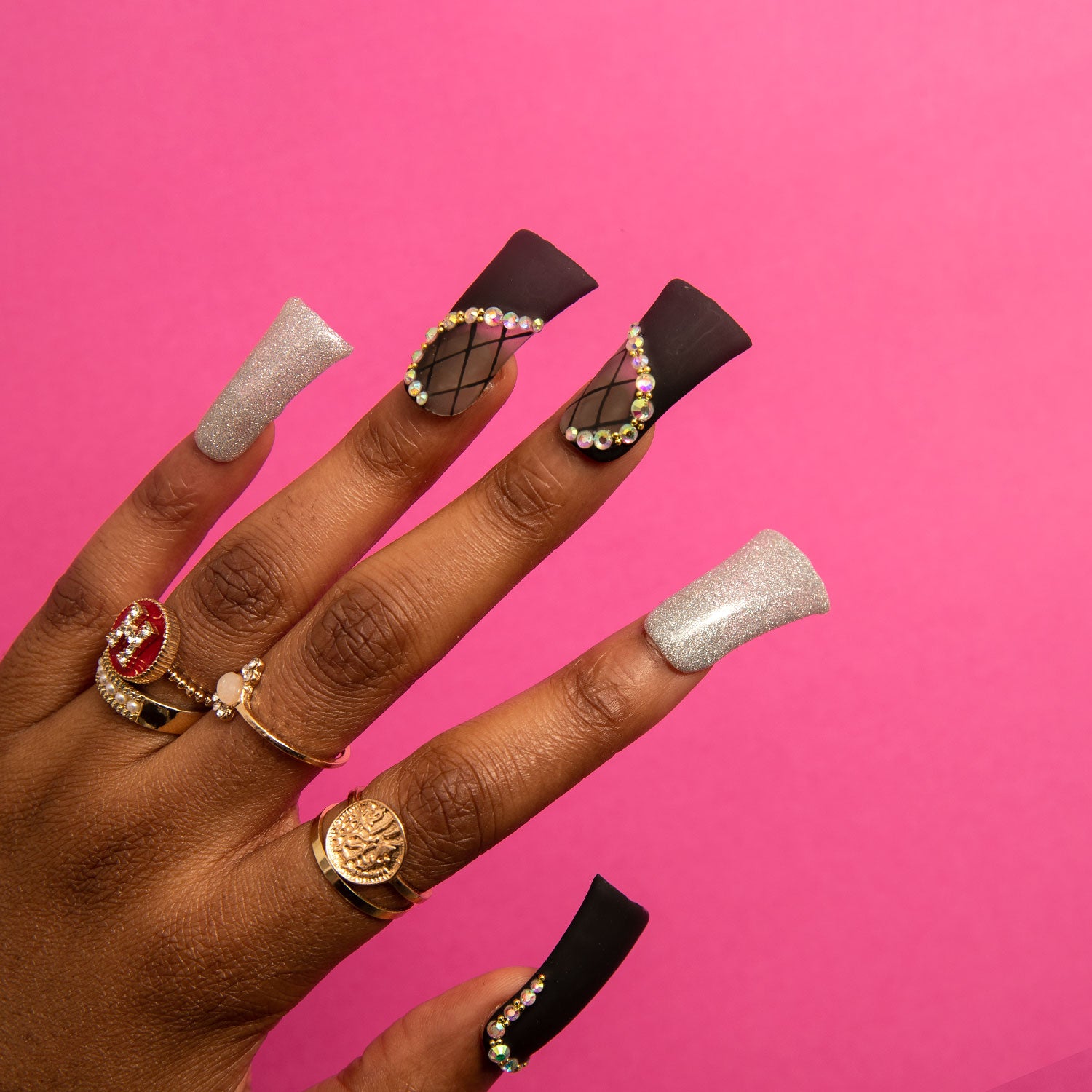 Hand with Lovful's Black Lace press-on nails on a pink background. The nails feature lace patterns, silver shimmer, and rhinestones, exuding elegance. Model's hand adorned with gold rings and gemstones.