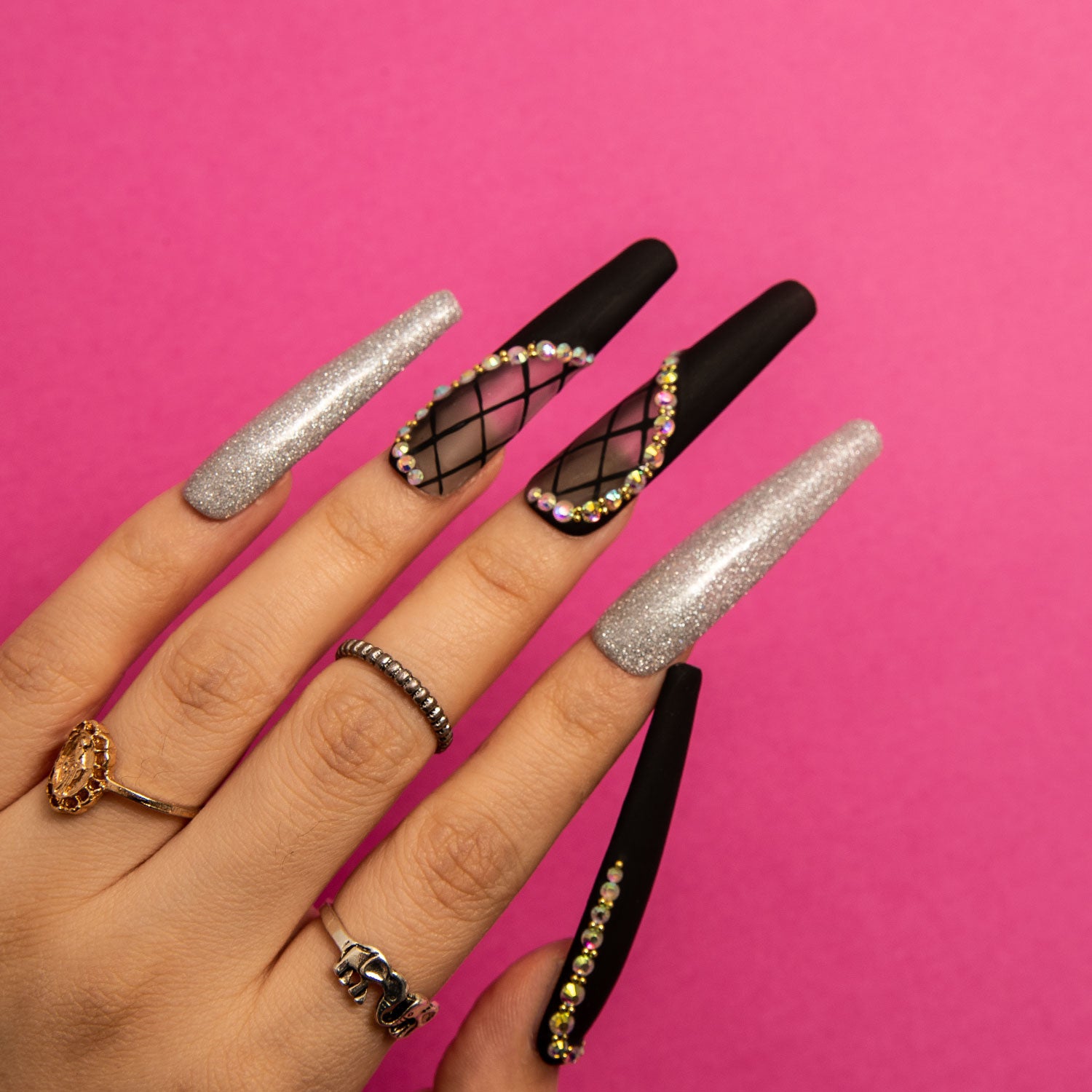 Hand with long coffin-shaped press-on nails featuring black lace with rhinestones and silver shimmer design on a pink background. Ideal for high-end events or romantic dinners.