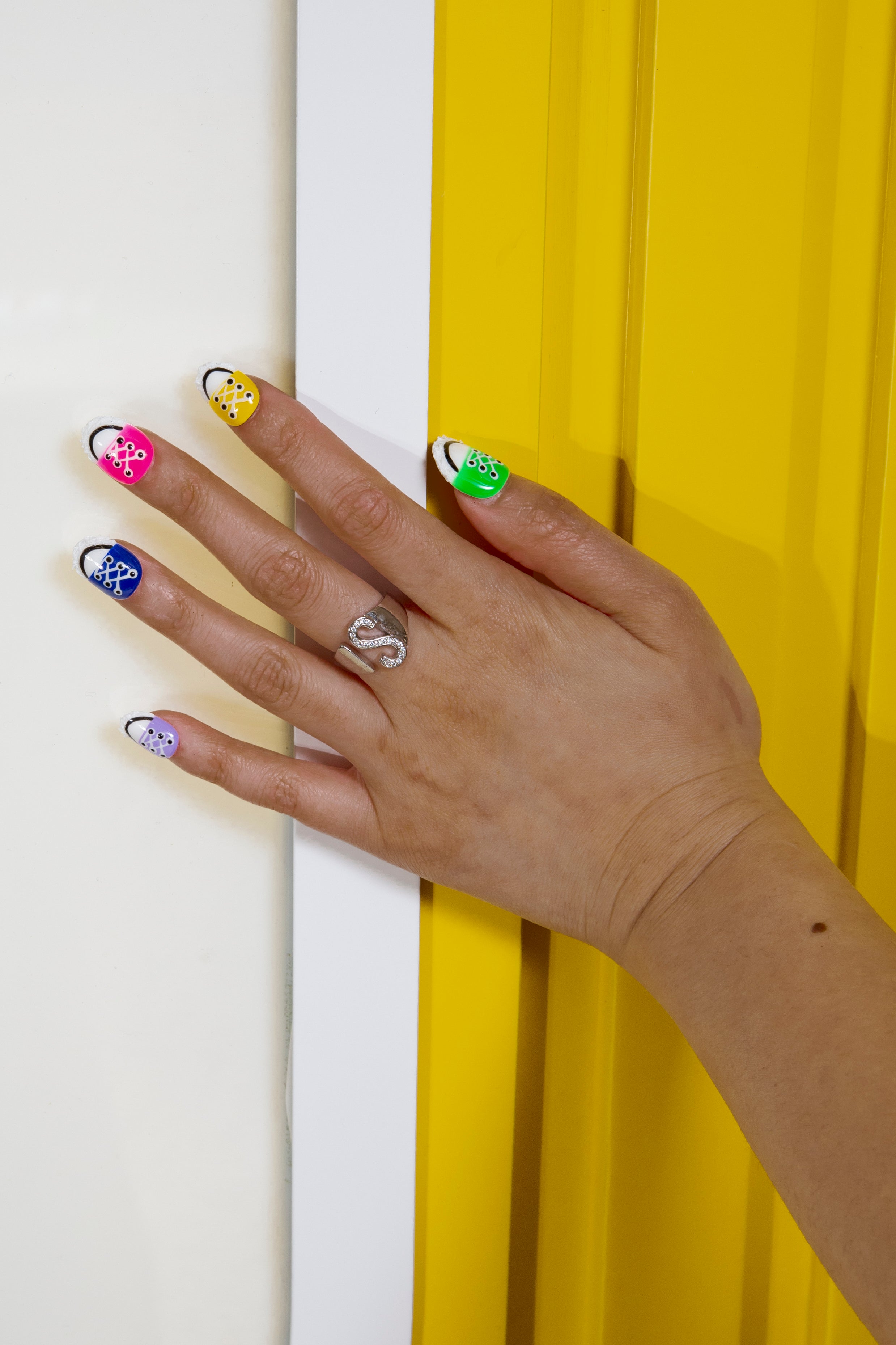 Hand with silver ring against a yellow background showcasing colorful canvas shoe-inspired press-on nails in purple, blue, red, yellow, and green from Lovful.com's 'Walk in Spring' collection.