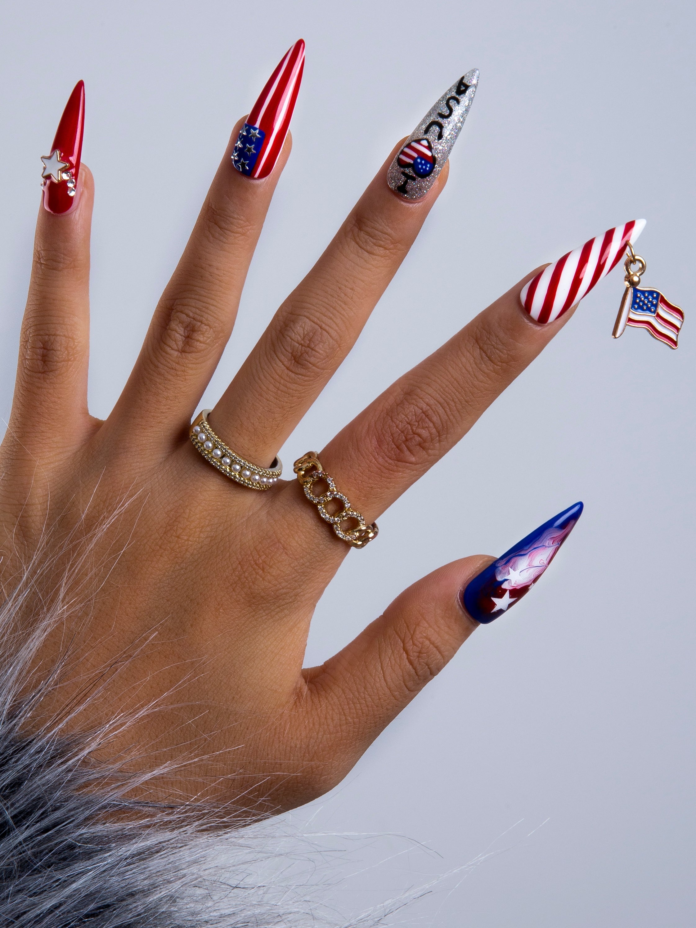 Hand featuring coffin-shaped press-on nails with American flag designs in red, white, and blue. One nail has a USA flag charm. Gold rings adorn the fingers, and a gray fur piece is visible on the wrist.