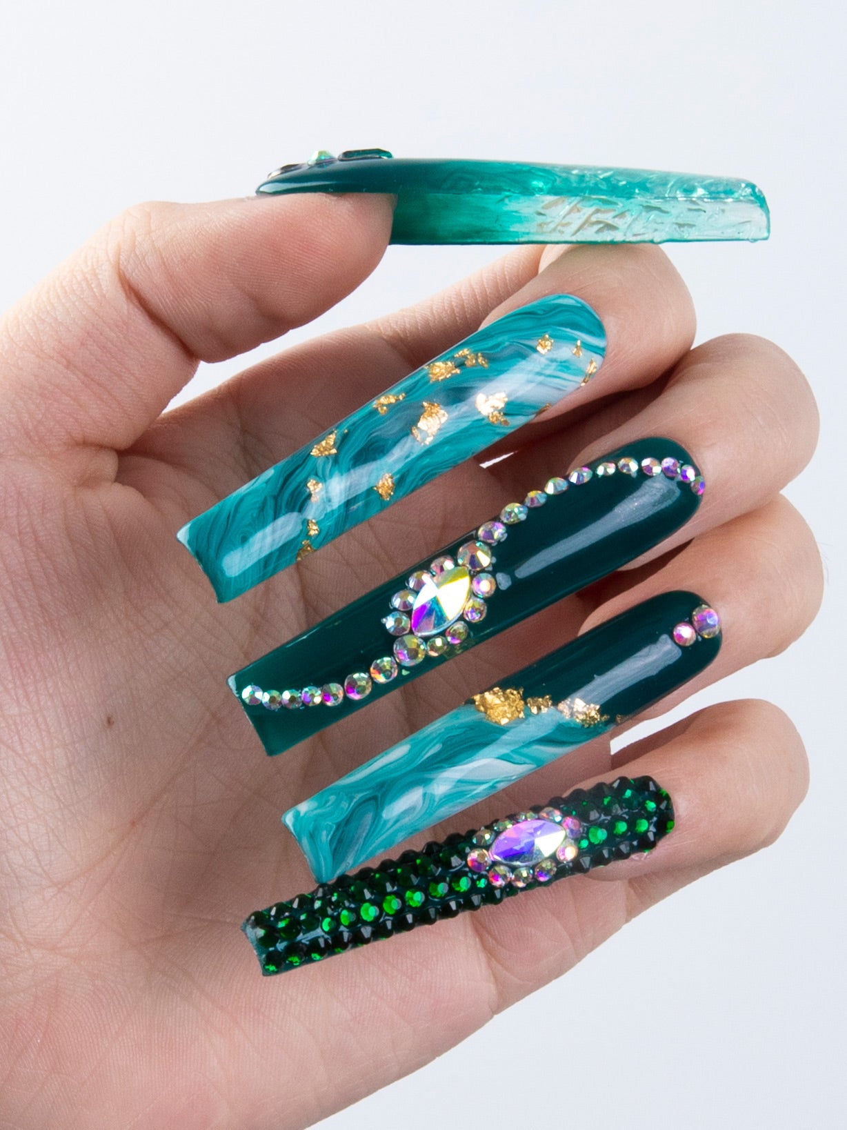 Hand showcasing the Emerald Envy press-on nails by Lovful.com, featuring intricate gold decor and crystal embellishments in vibrant shades of green. Long square shapes enhance the trendy design.