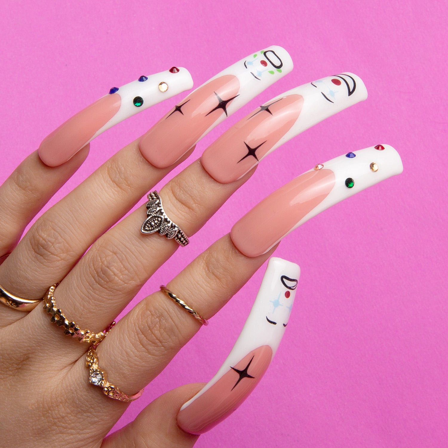 Hand with long, curved, circus clown-themed press-on nails, combining classic French tip design with clown faces and colorful rhinestones, against a pink background.