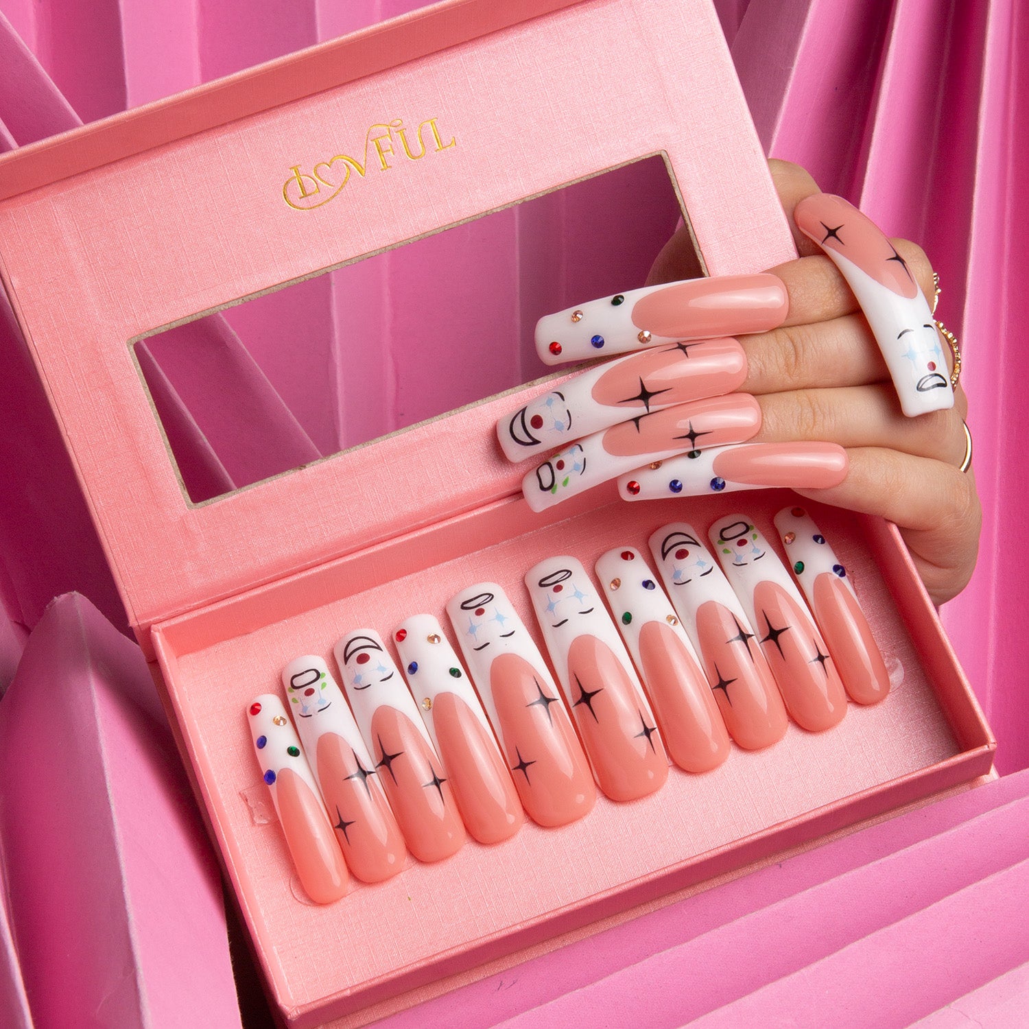 Circus Clown-themed long press-on nails by Lovful, with playful clown faces, polka dots, and cross designs, displayed in a pink box with a hand showcasing the applied nails.