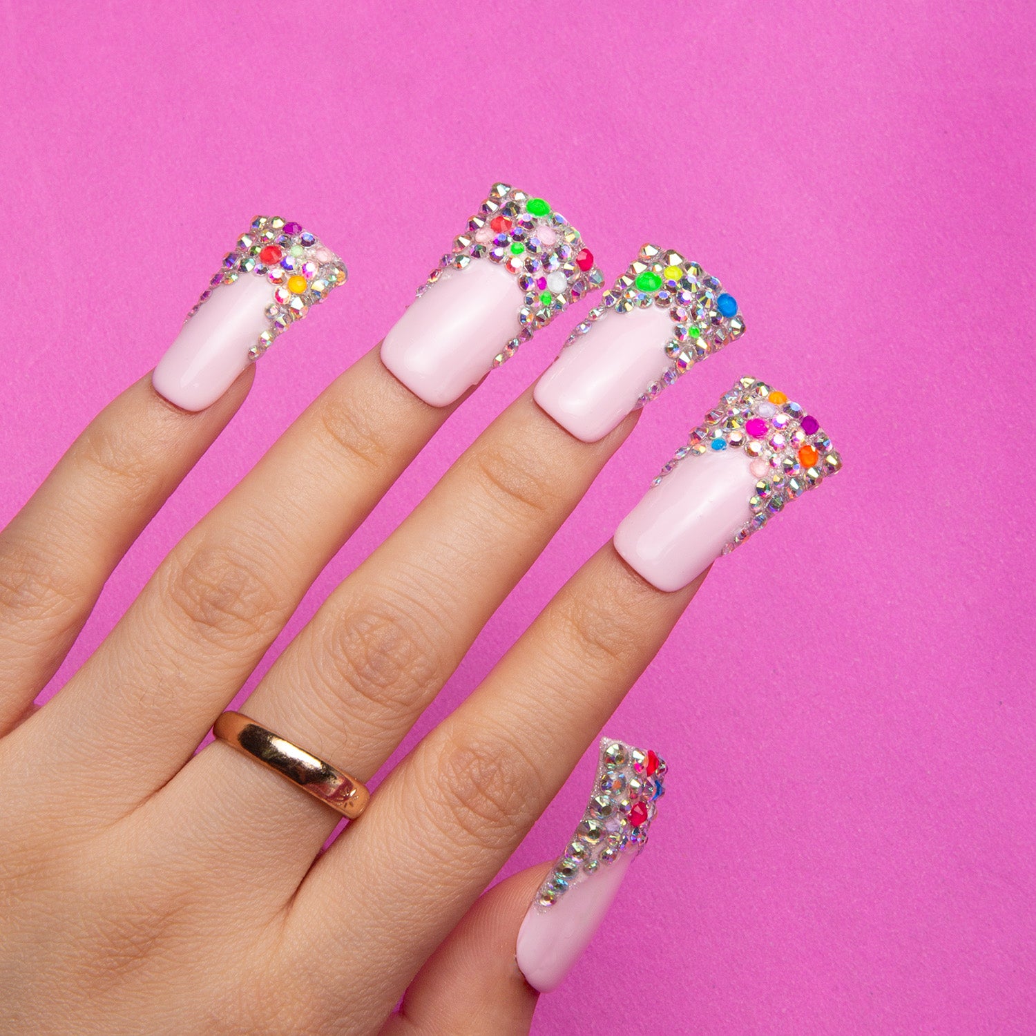 Hand with long press-on acrylic nails featuring a chic nude base and jelly bean design with colorful tiny beans on the tips against a pink background.