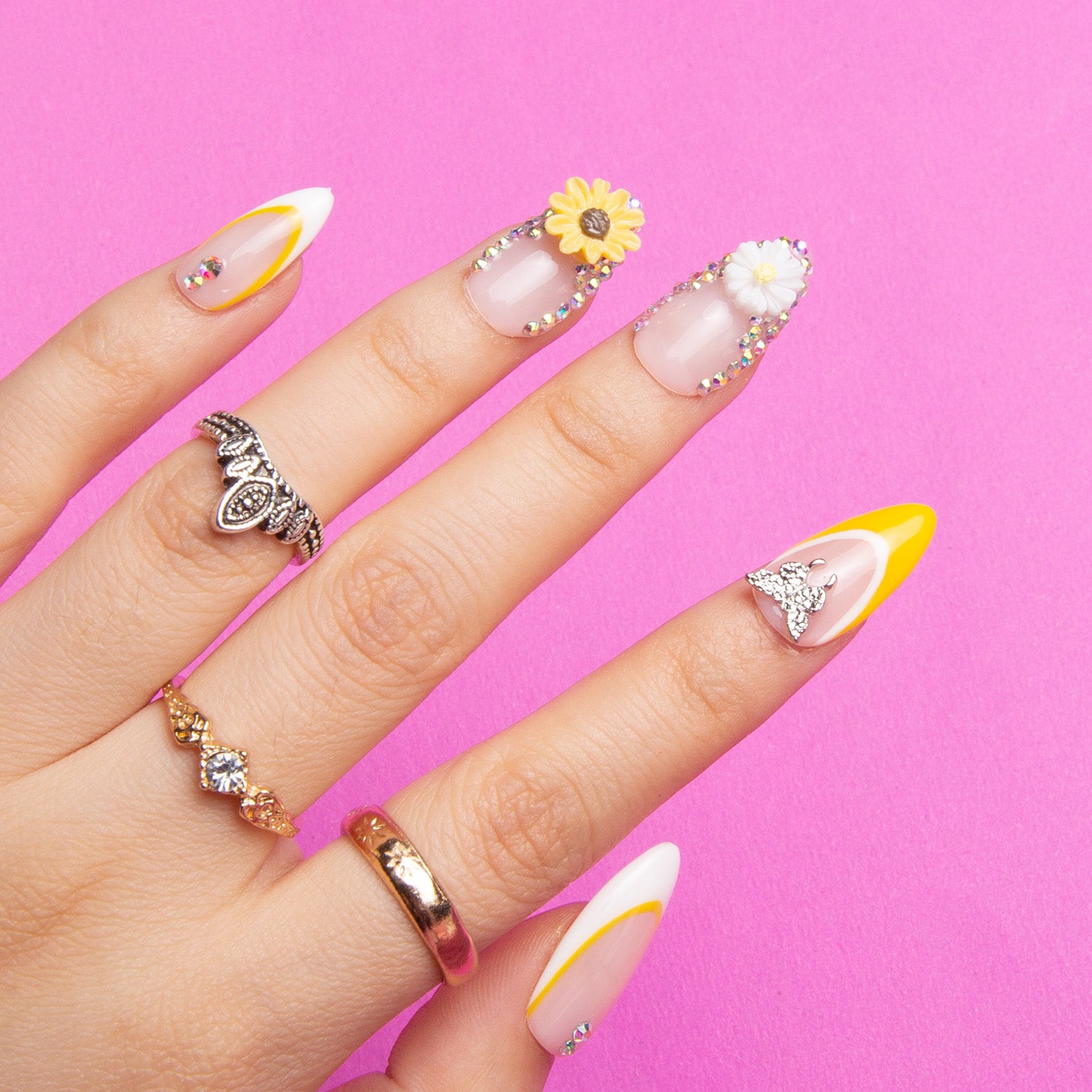 Close-up of Sunflower press-on nails with vibrant yellow French tips, white decor, and sunflower designs on almond-shaped nails, complemented by silver and gold rings, set against a pink background.