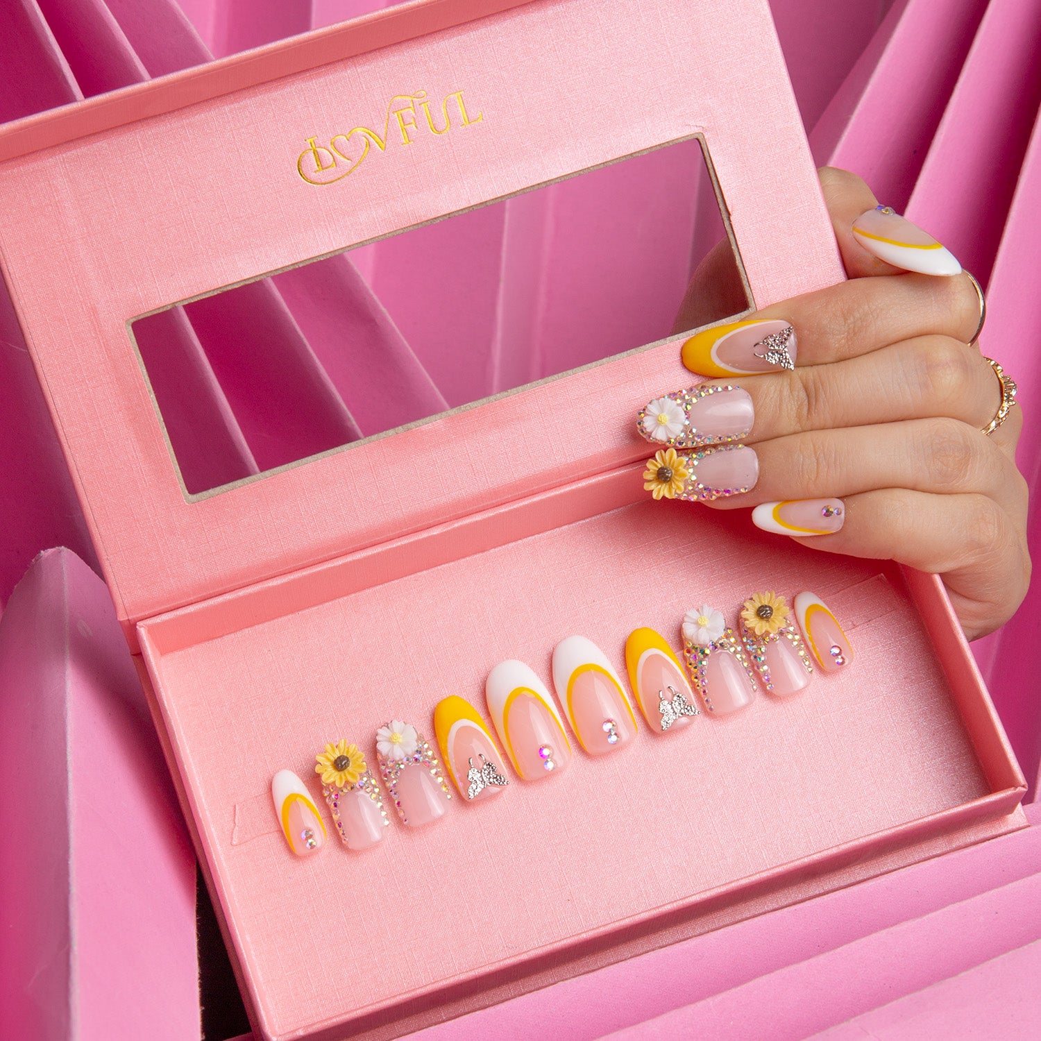 Boxed set of Lovful 'Sunflower' press-on nails with yellow and white French tips adorned with sunflowers, displayed by a hand with matching nails against a pink background.