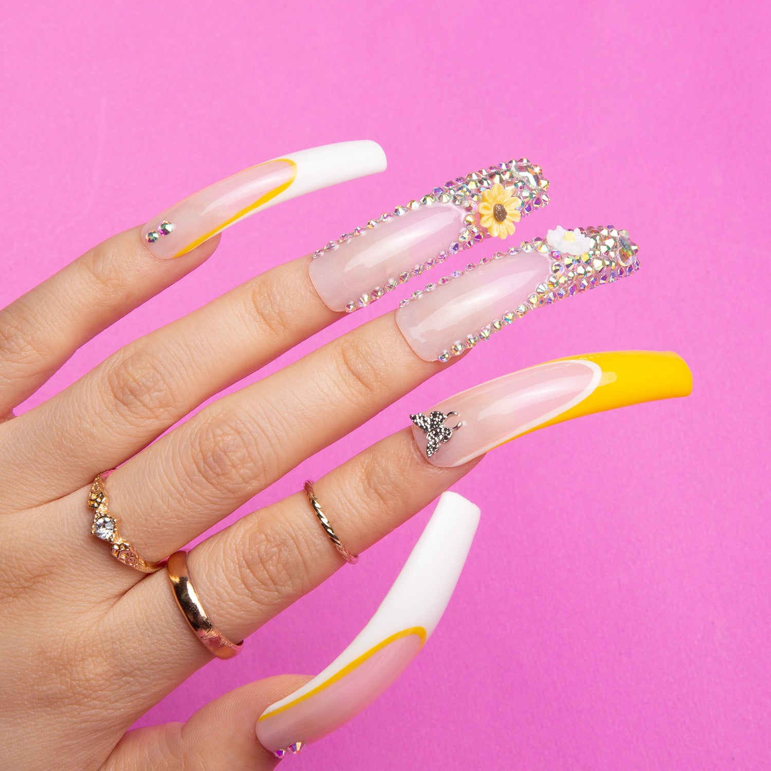 Hand with 'Sunflower' press-on acrylic nails, featuring yellow and white French tips decorated with sunflower decals and rhinestones, on a pink background