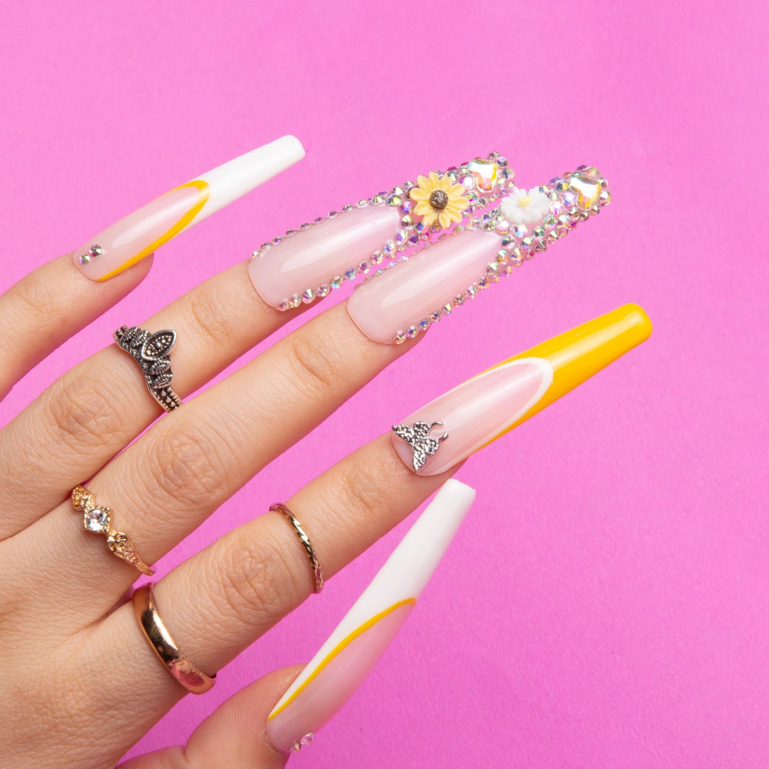 Hand with long coffin-shaped 'Sunflower' press-on nails, featuring white French tips, yellow accents, sunflower decor, and rhinestones on a bright pink background. The hand is wearing rings.