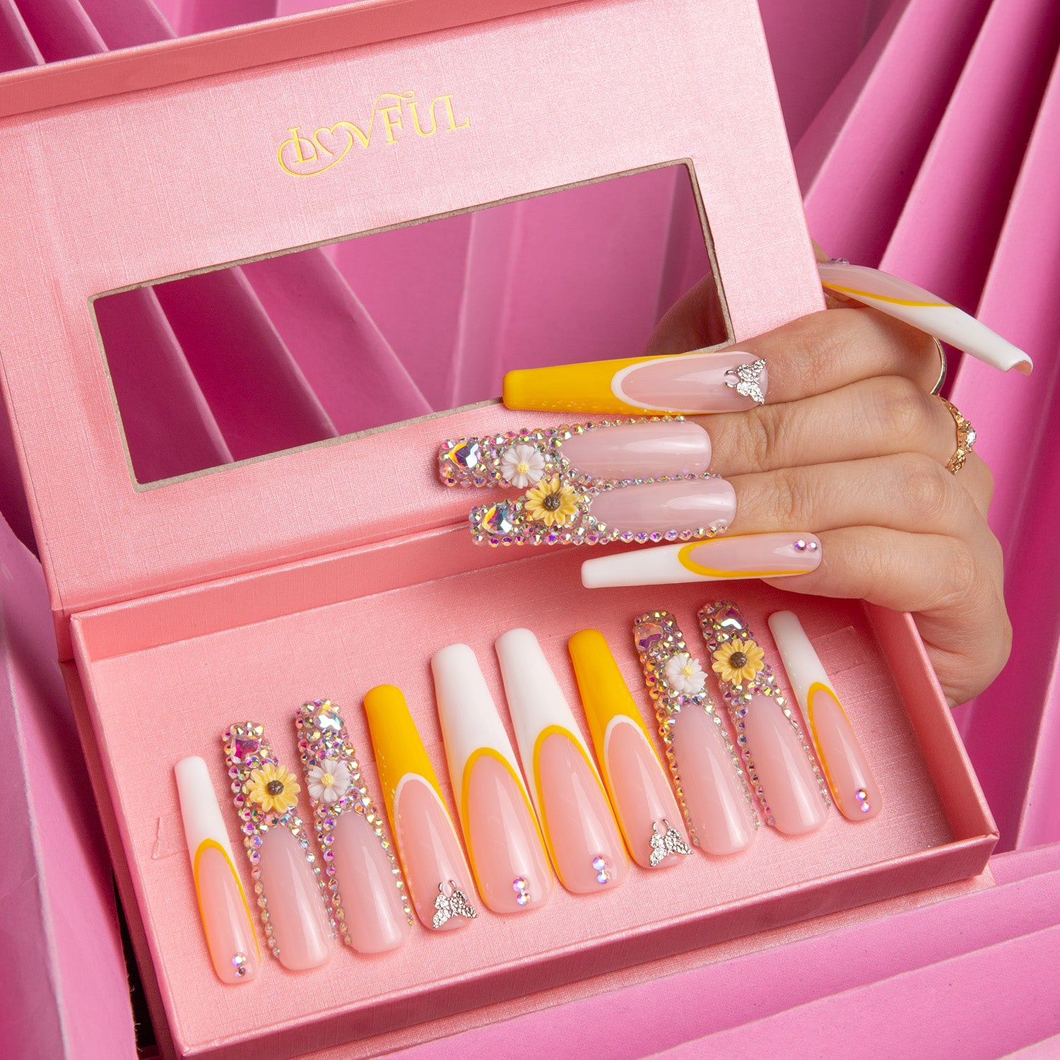 Sunflower press-on nails in open pink box, featuring yellow hues, white French tips, and sunflower designs. A hand is shown wearing the nails.