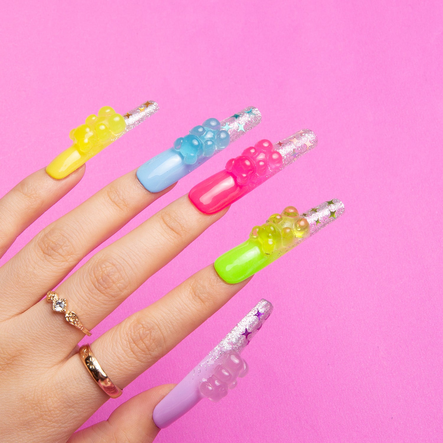 Hand with colorful press-on coffin nails in yellow, blue, pink, green, and purple with glowing gummy bear charms, set against a pink background.