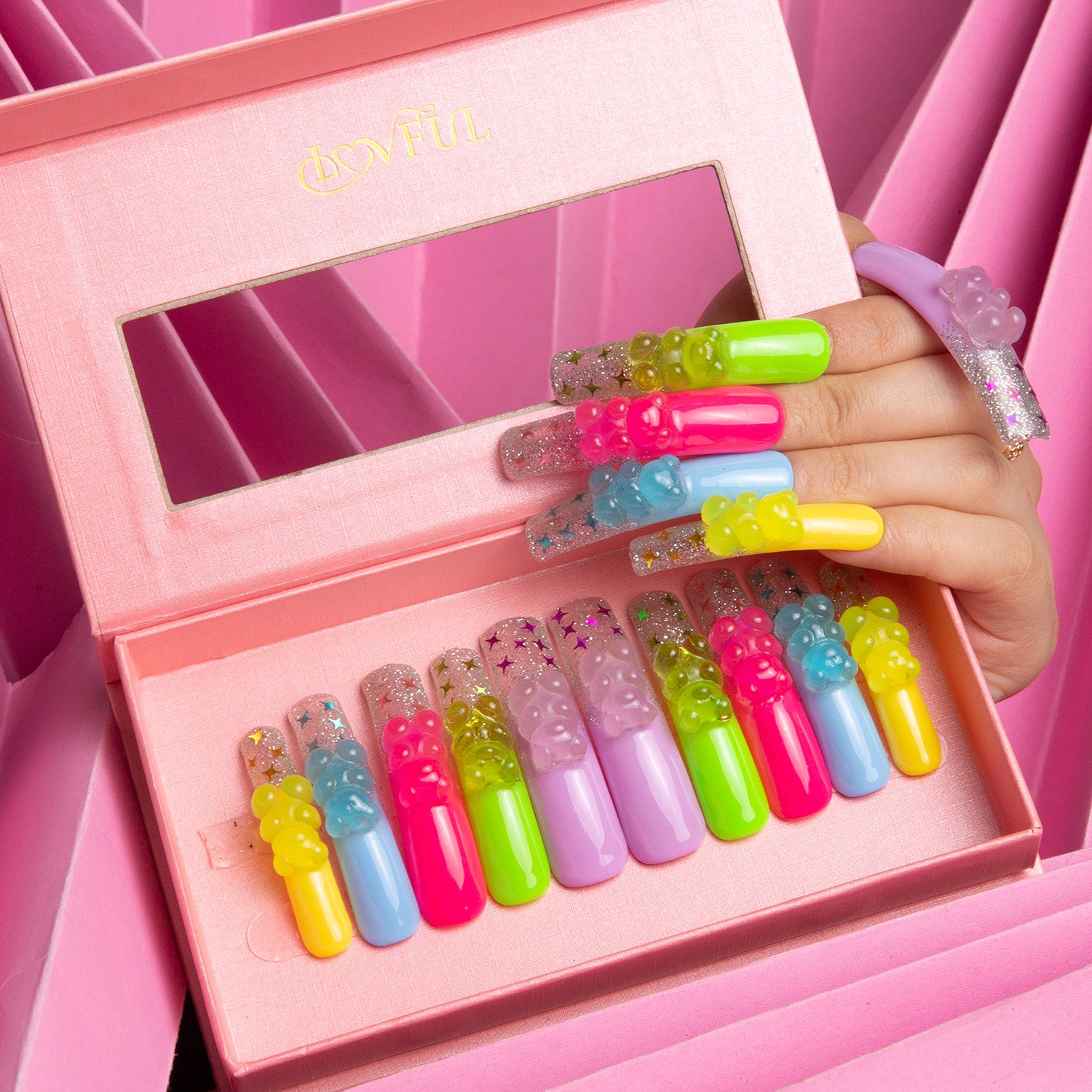 Lovful 'Sweet Wonderland' press-on nails in a pink box with mirror. Nails feature vibrant colors including candy pink and sunshine yellow, with glow-in-the-dark gummy bear charms, held by a hand.