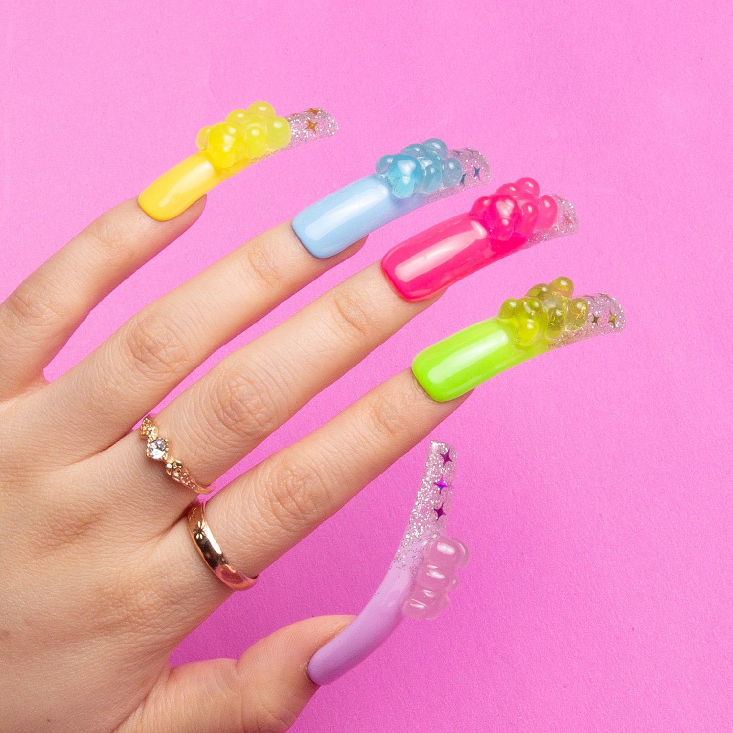 Hand with long, colorful 'Sweet Wonderland' press-on acrylic nails by Lovful.com, featuring yellow, blue, pink, green, and purple nails with glowing gummy bear charms on a pink background.