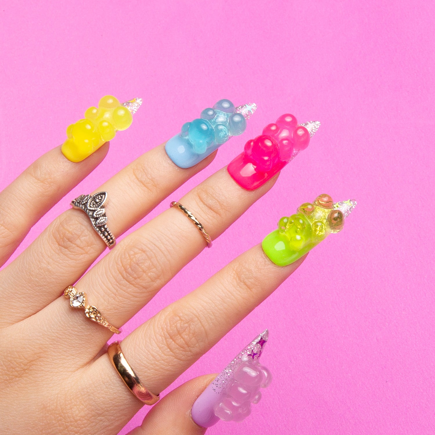 Close-up of a hand with 'Sweet Wonderland' press-on nails by Lovful. Each nail is a different vibrant color and decorated with glow-in-the-dark gummy bear charms. The hand is wearing multiple rings, against a pink background.