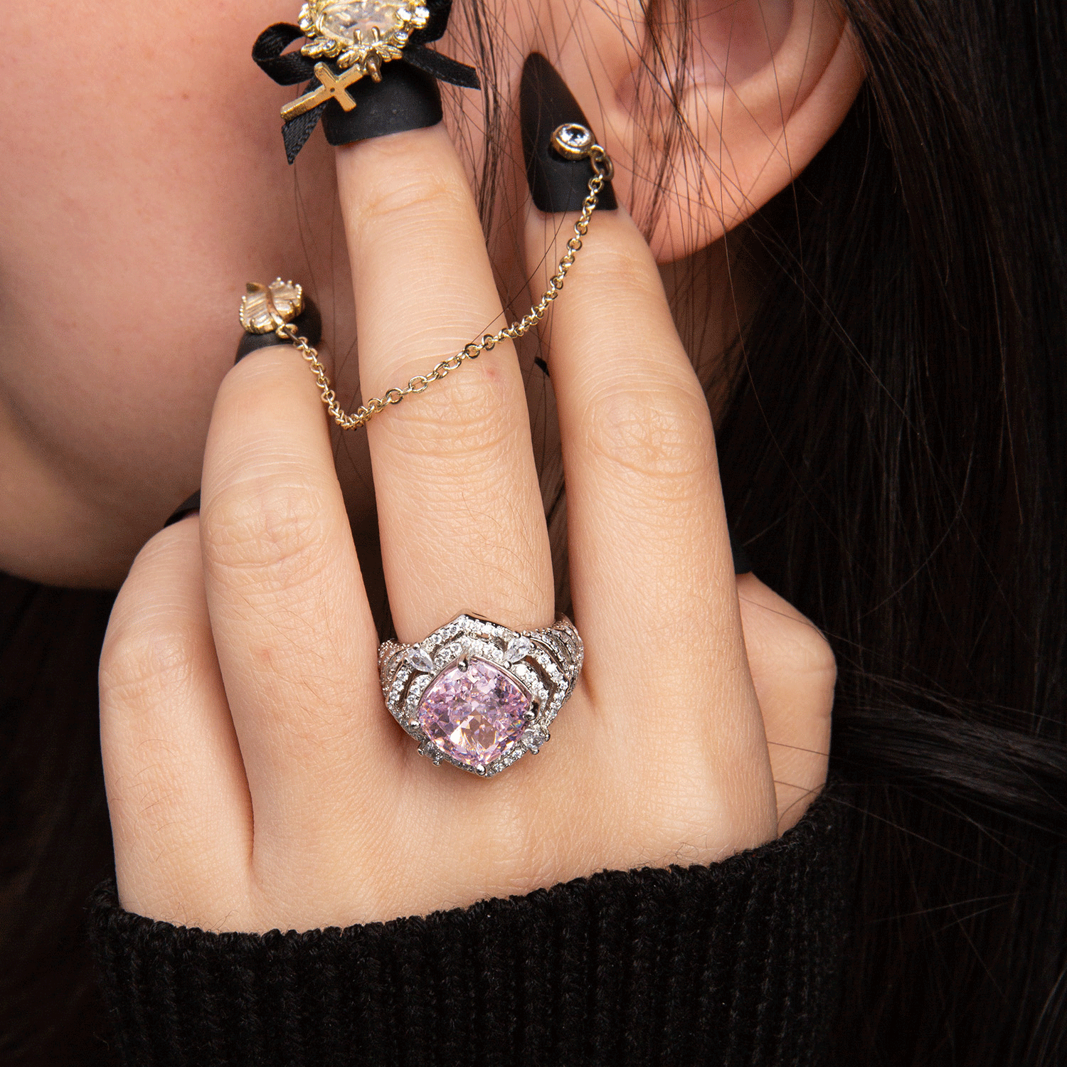 Silver ring with a large pink gemstone and diamond accents on ring finger, black nail polish, and black finger accessory with chain and gold cross.