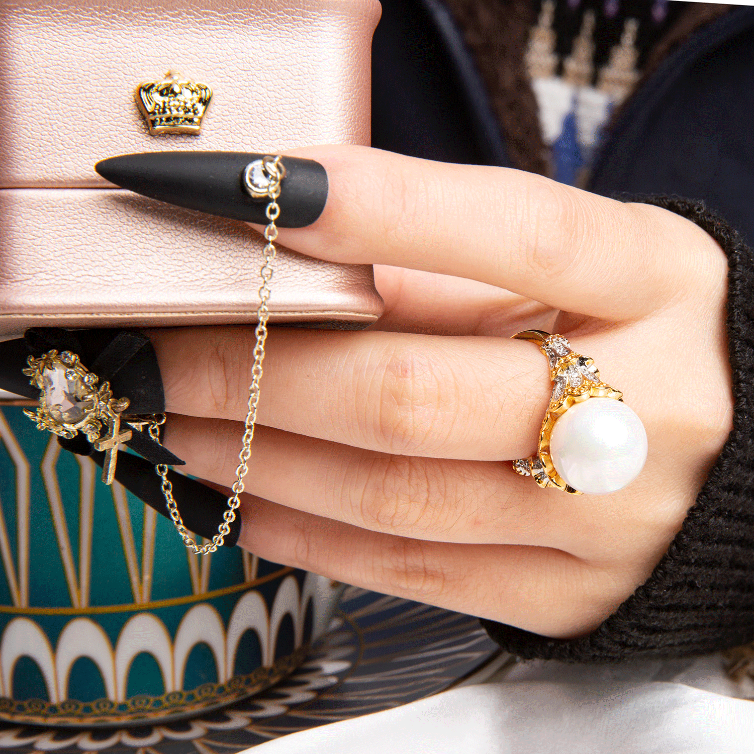 Hand adorned with fashionable adjustable rings, one with a large pearl and gold setting, another with a chain connected to black matte press-on acrylic nail with crown detail, on a pink jewelry box background.