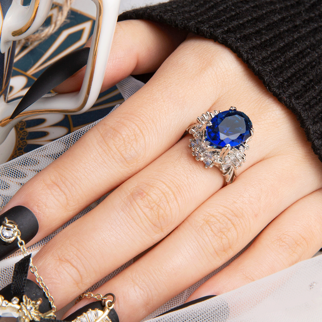 Hand wearing a silver adjustable ring with a large blue oval gemstone and intricate details, complemented by black acrylic nails with gold accents and rhinestones.