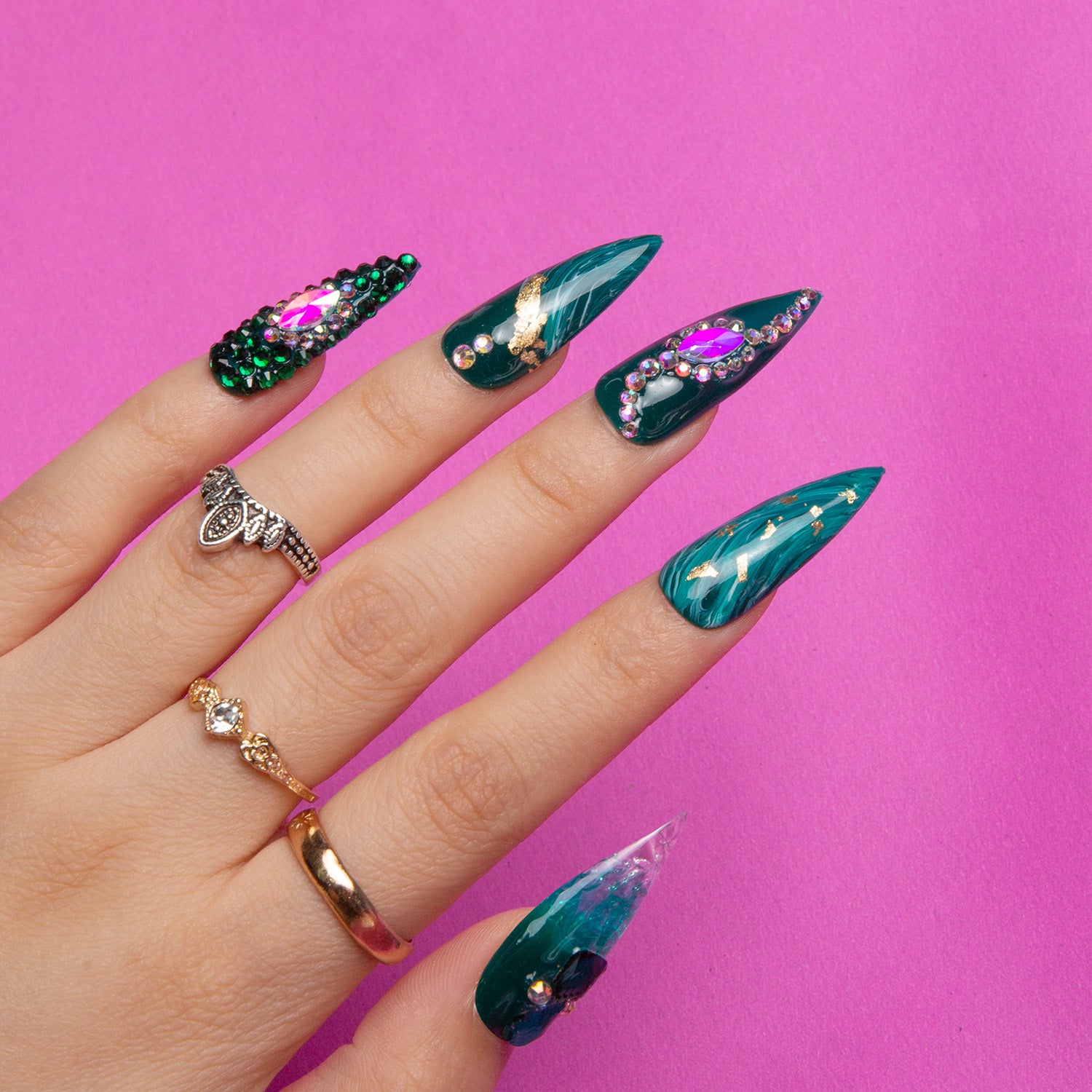 Hand with Emerald Envy Stiletto Press-On Nails featuring green hues, gold decor, and jewel embellishments, set against a bright pink background.
