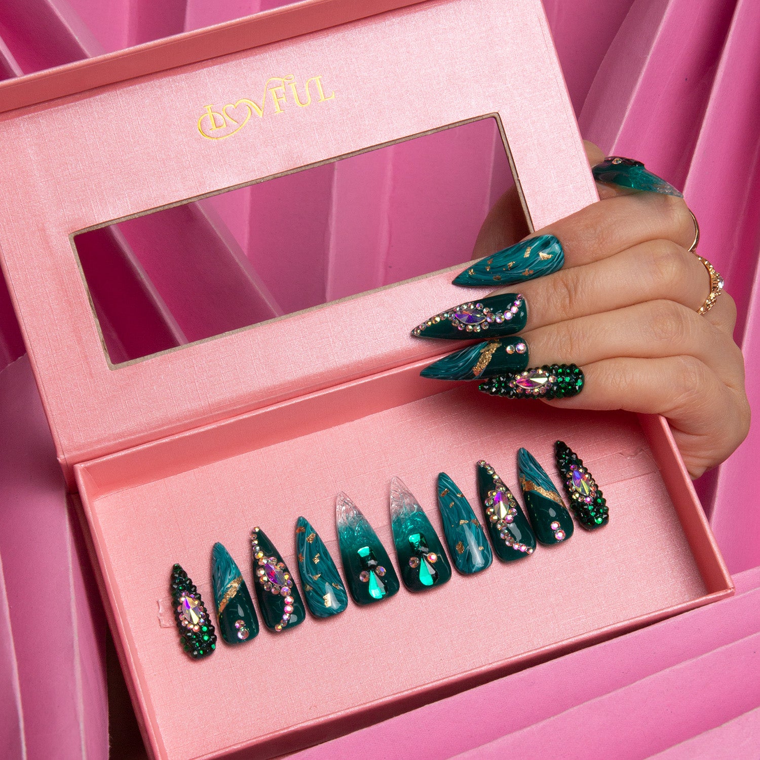Hand wearing long, stiletto-shaped Emerald Envy press-on nails with intricate green and gold designs, displayed in a pink Lovful branded box against a pink textured background.