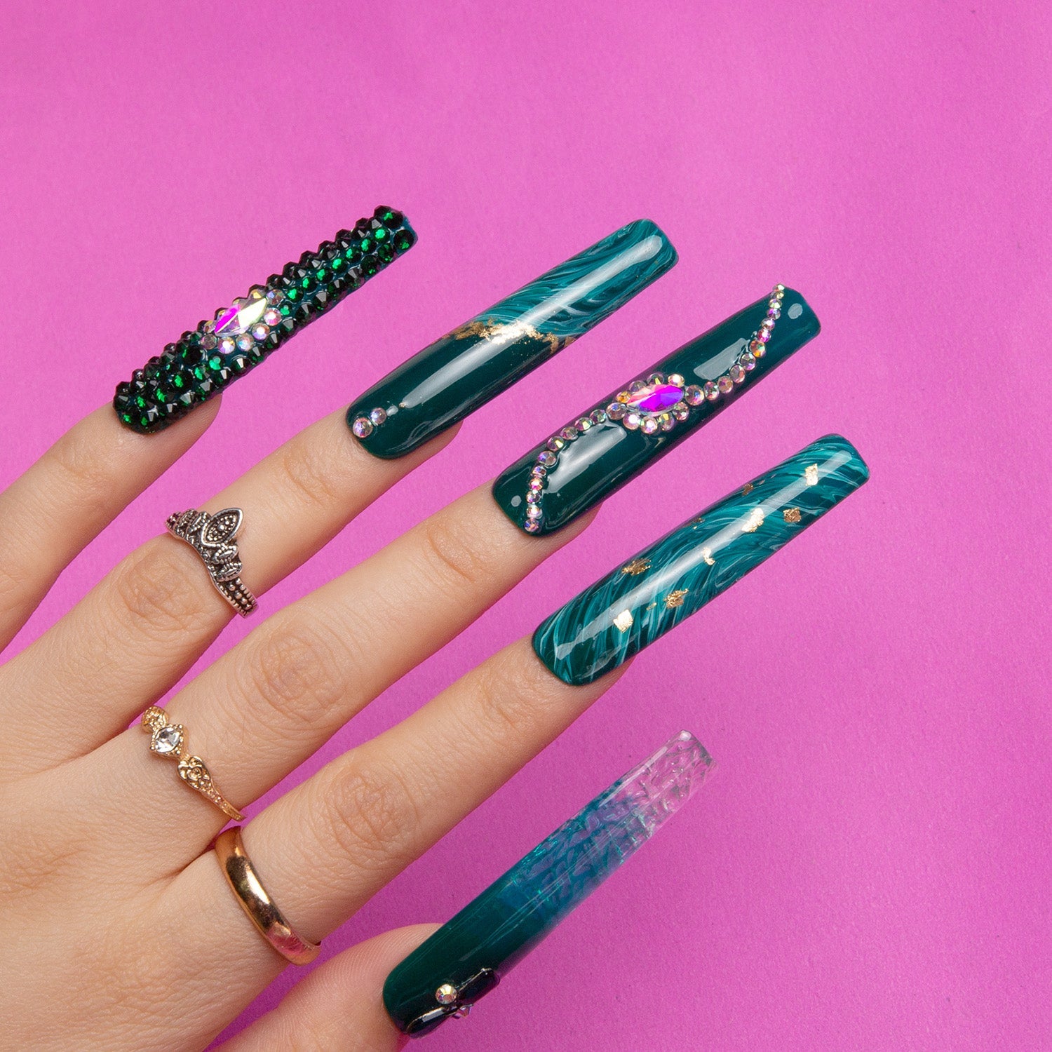 Hand with square-shaped press-on acrylic nails in emerald green with gold decor, gemstones, and liquid flowing design, set against a pink background.