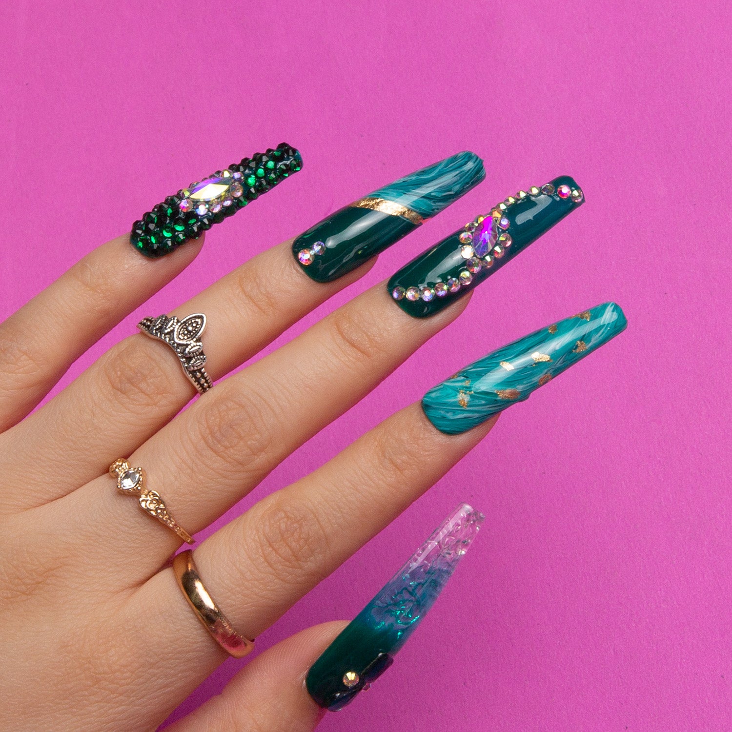 Hand wearing Emerald Envy coffin-shaped press-on nails decorated with intricate gold accents, green shades, and gemstones evoking a lush woodland aesthetic against a pink background.