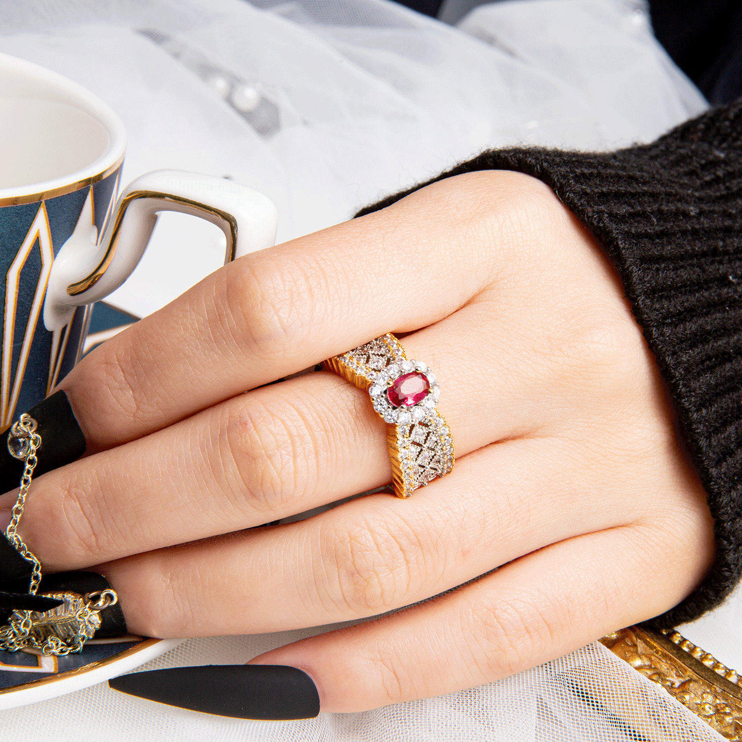 Hand with black pressed-on nails wearing a stylish gold ring with red gemstone, holding a teacup.