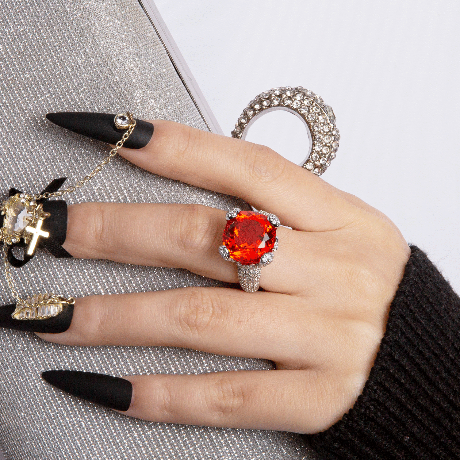 Fashionable hand with black matte press-on nails wearing a large red gemstone ring, holding a silver clutch bag