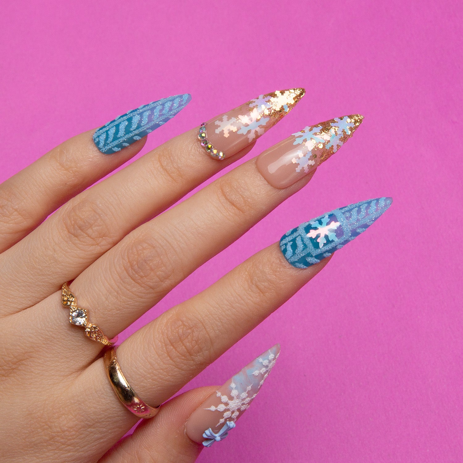 Hand with 'Winter Sunshine' press-on nails in stiletto shape, blue to clear gradient design, adorned with white snowflakes and gold glitter tips, displayed on a bright pink background.