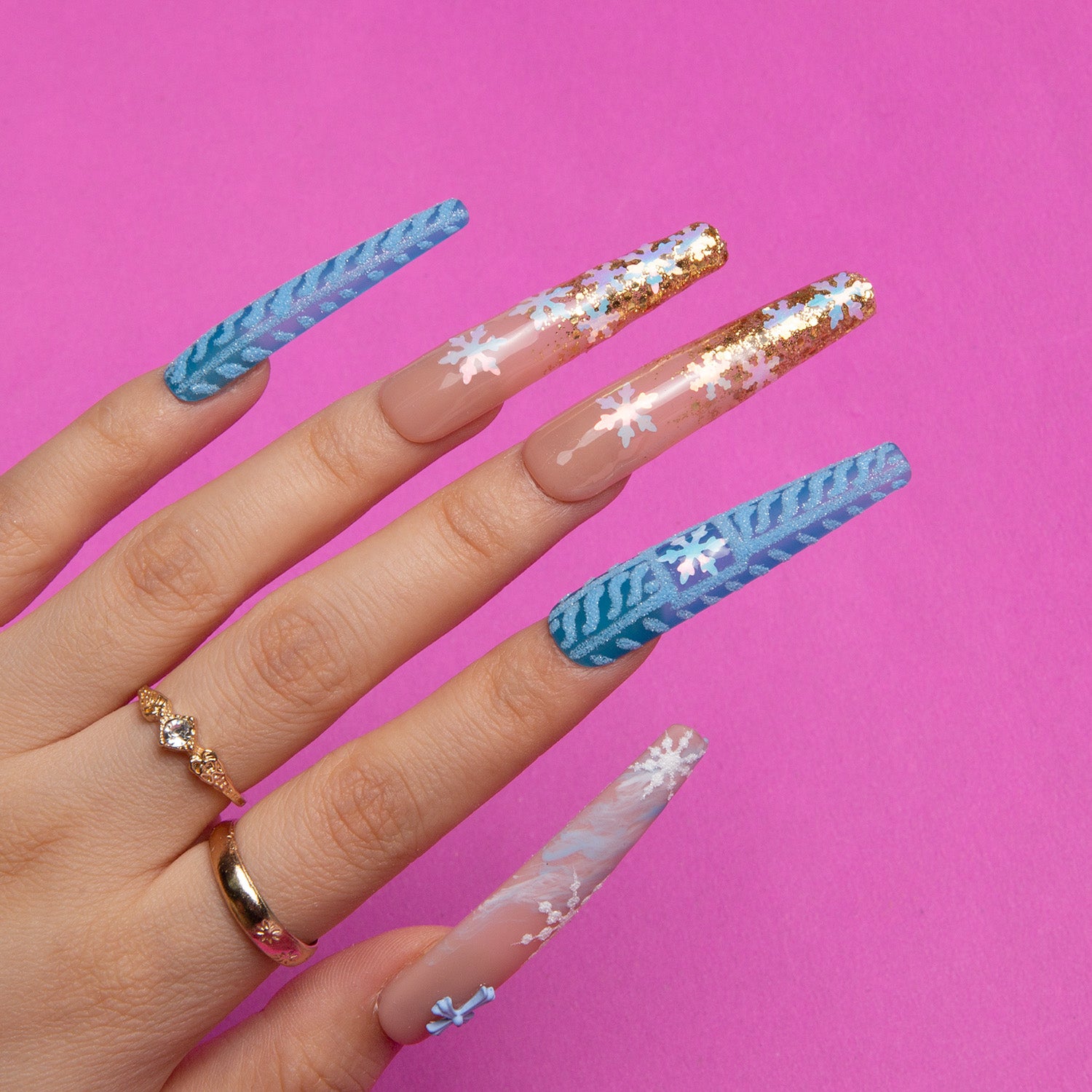 Hand displaying Winter Sunshine coffin-shaped press-on nails with blue hues, snowflakes, and golden glitter, against a bright pink background, featuring a gold ring with gemstones.