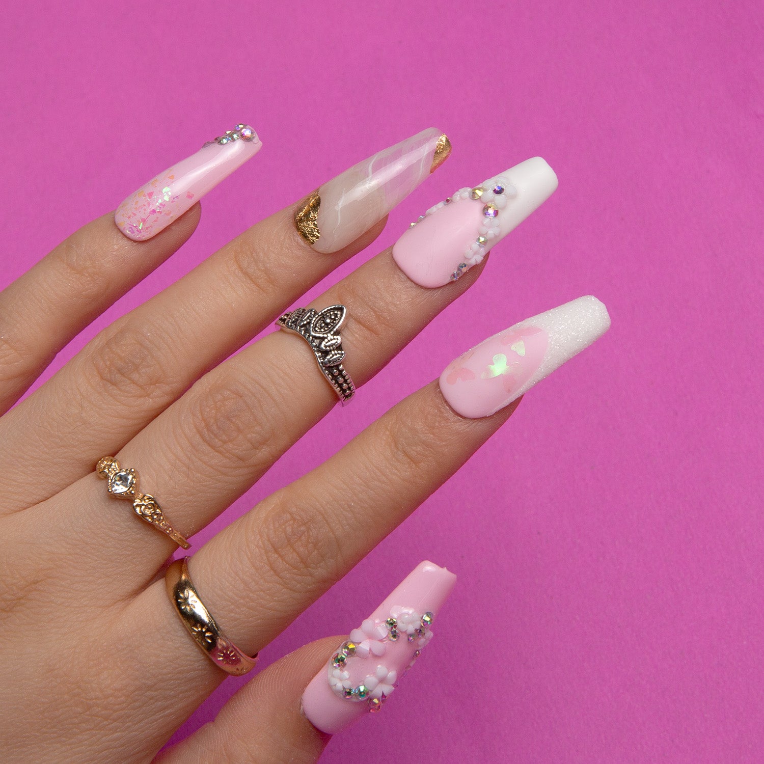 Hand with press-on acrylic nails in light pink and white featuring delicate French tips, heart-shaped flower designs, and crystal embellishments, wearing multiple elegant rings, against a pink background. Ideal for weddings.