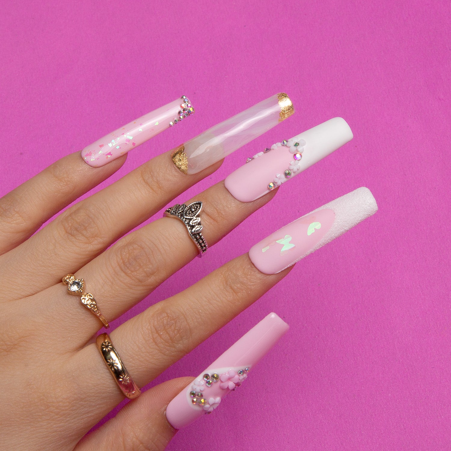 Hand showcasing long square press-on nails in light pink and white with intricate heart-shaped flower and gem designs on a pink background, worn with elegant rings