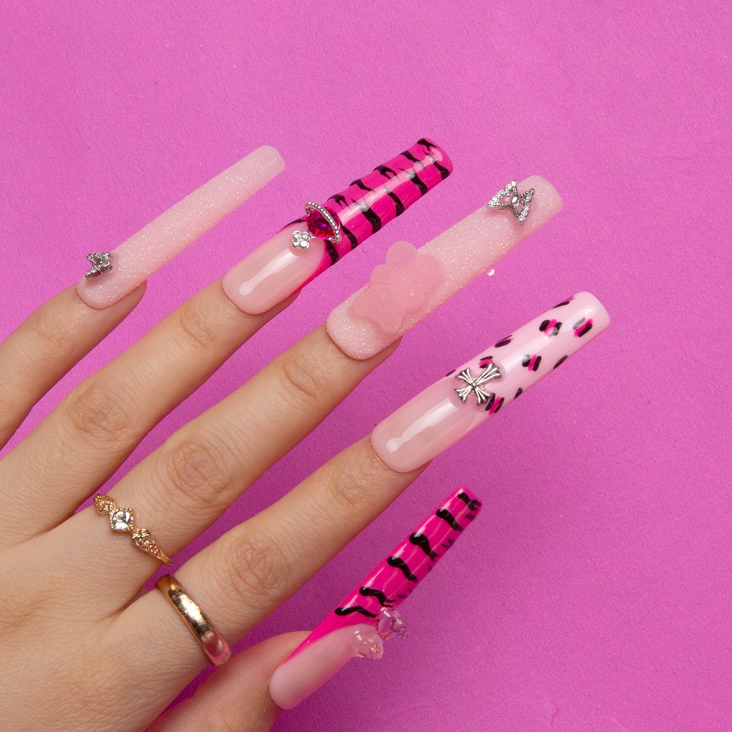 Hand displaying 'Safari Love' press-on nails with hot pink French tips, black leopard prints, glitter accents, and butterfly and bow charms on a pink background.