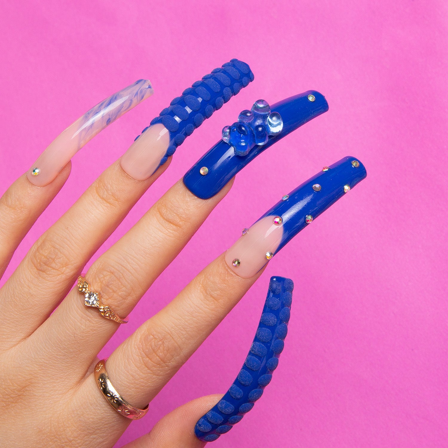 Hand displaying 'Little Blue Bear' rhinestone French tip press-on nails in curve shape, featuring blue gummy bears, shiny rhinestones, and hand-drawn 3D dots on a pink background.