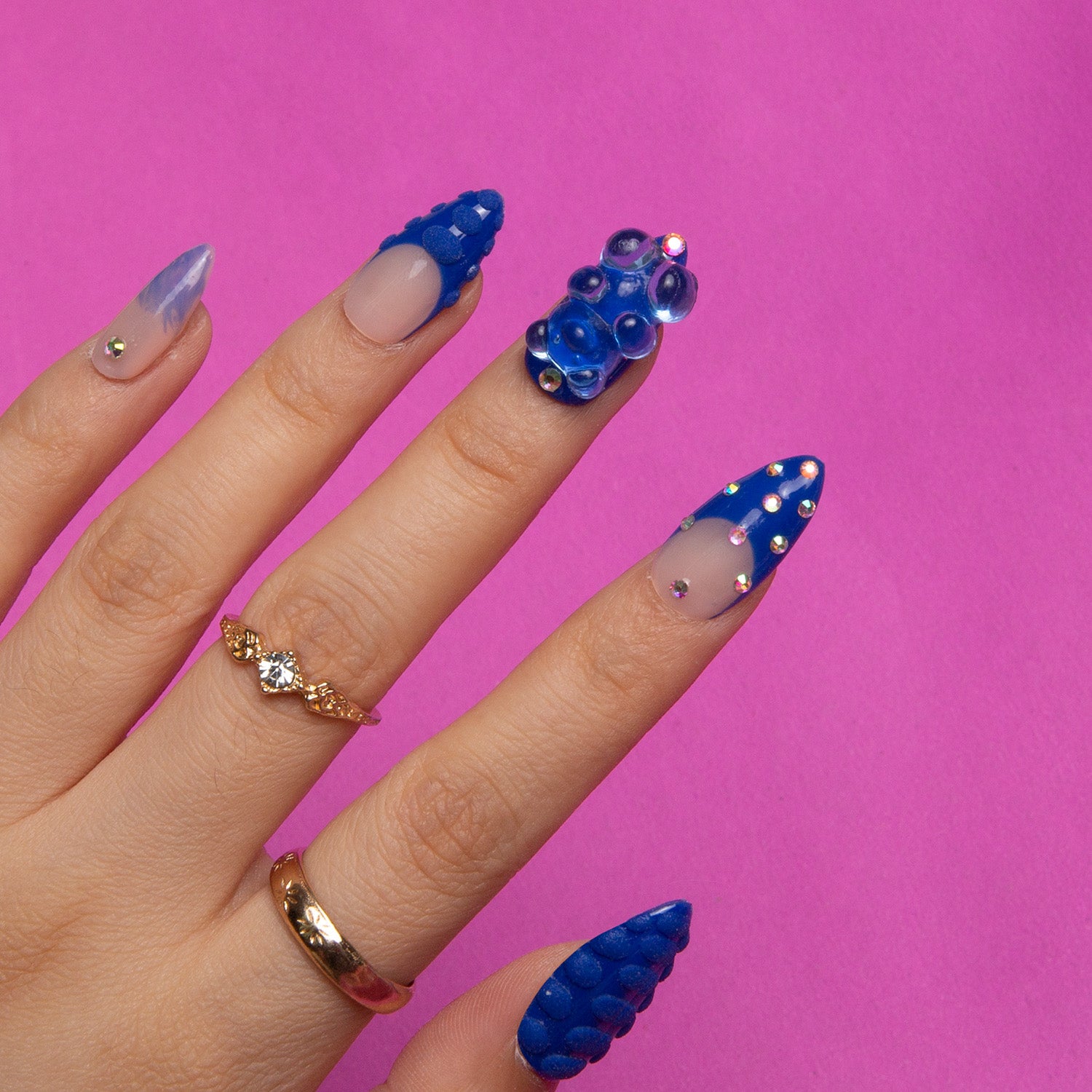 Hand with almond-shaped press-on nails featuring blue rhinestones, gummy bears, and 3D dots on a pink background, complemented by gold rings.