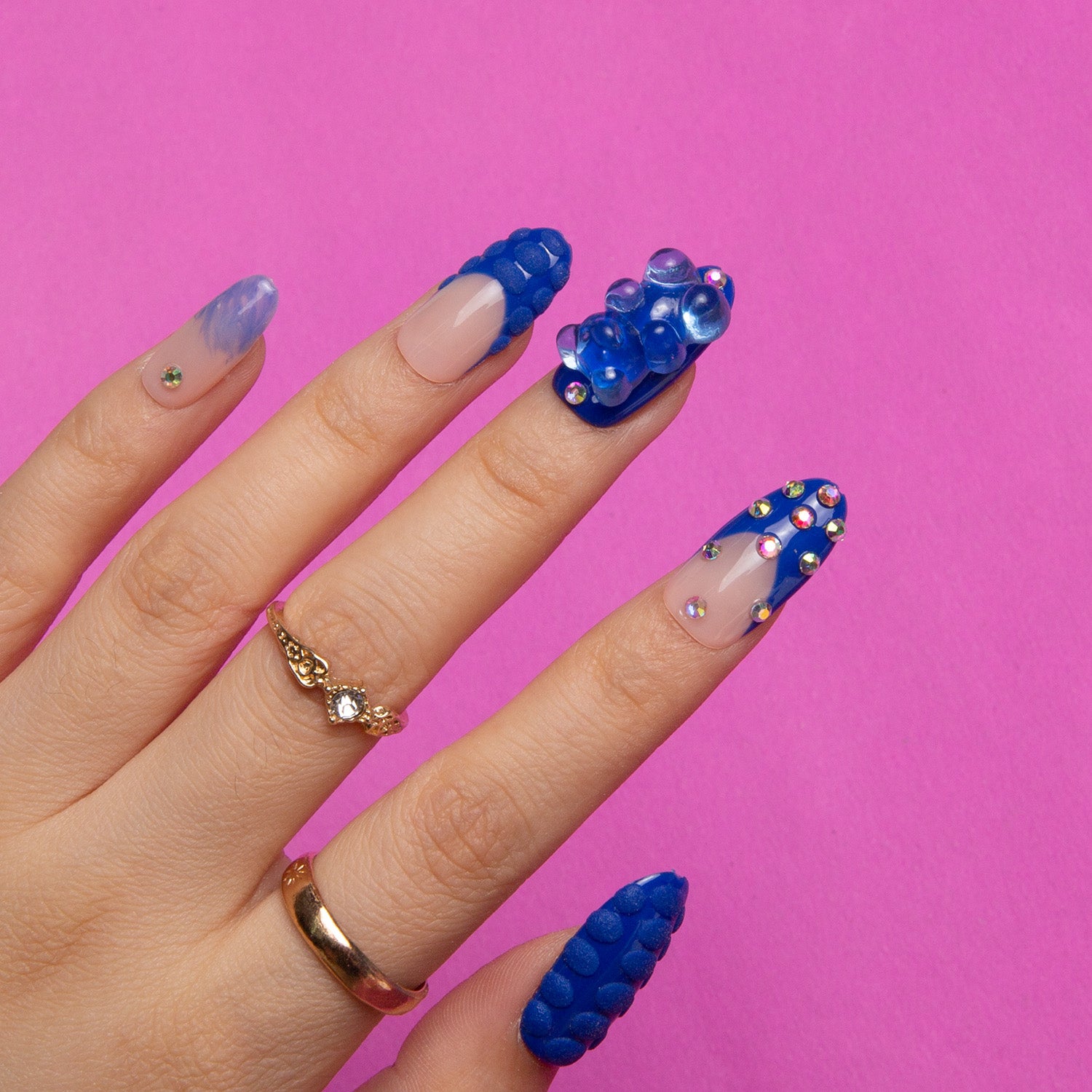 Hand with Little Blue Bear Rhinestone French tip nails featuring blue hue, gummy bear designs, rhinestones, and 3D dots, against a pink background. Fingers adorned with gold rings.