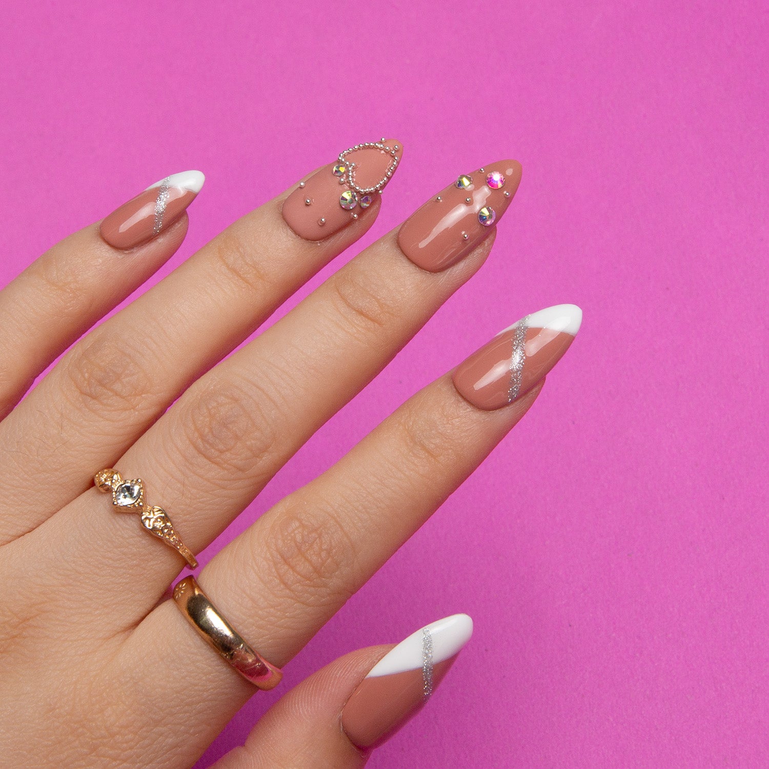 Almond shaped press-on nails with nude base and white French tips, adorned with silver glitter, rhinestones, and golden chain design, displayed on a hand against a pink background. Gold ring adds elegance.