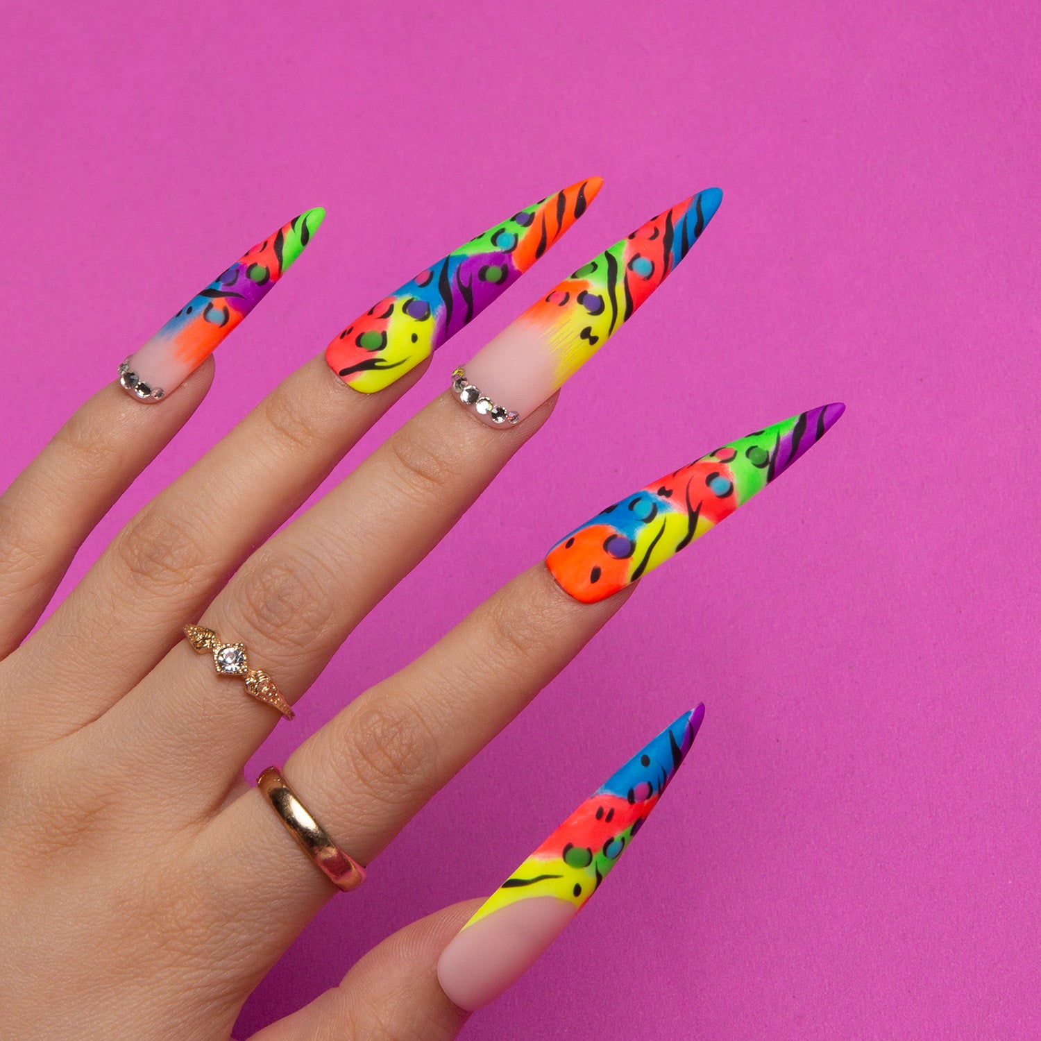 Long stiletto-shaped press-on nails with colorful bright French tips and leopard prints on a hand against a pink background, adorned with rings.