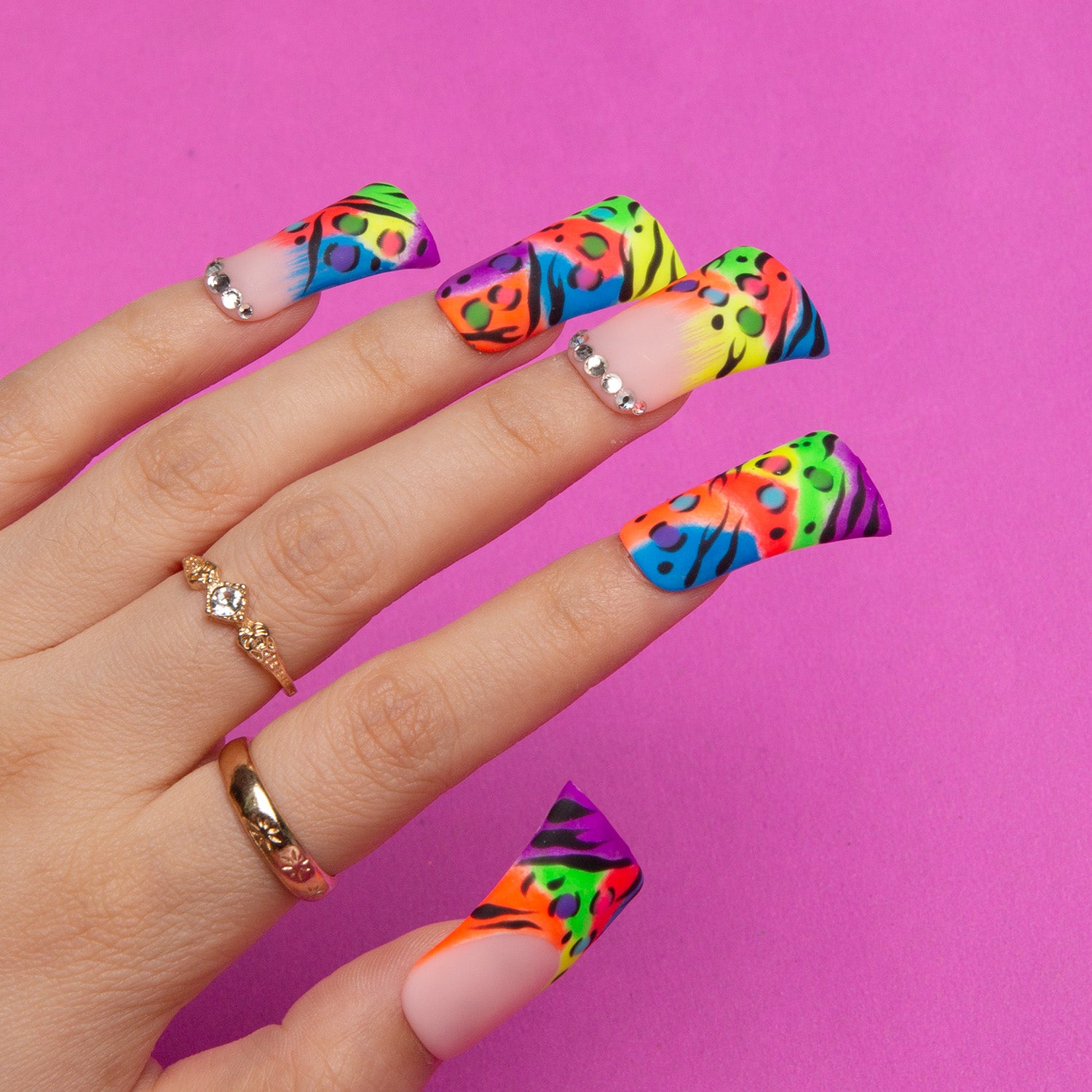 Hand with press-on duck-shaped nails featuring bright French tips with leopard prints on a pink background, complemented by gold rings with small diamonds.