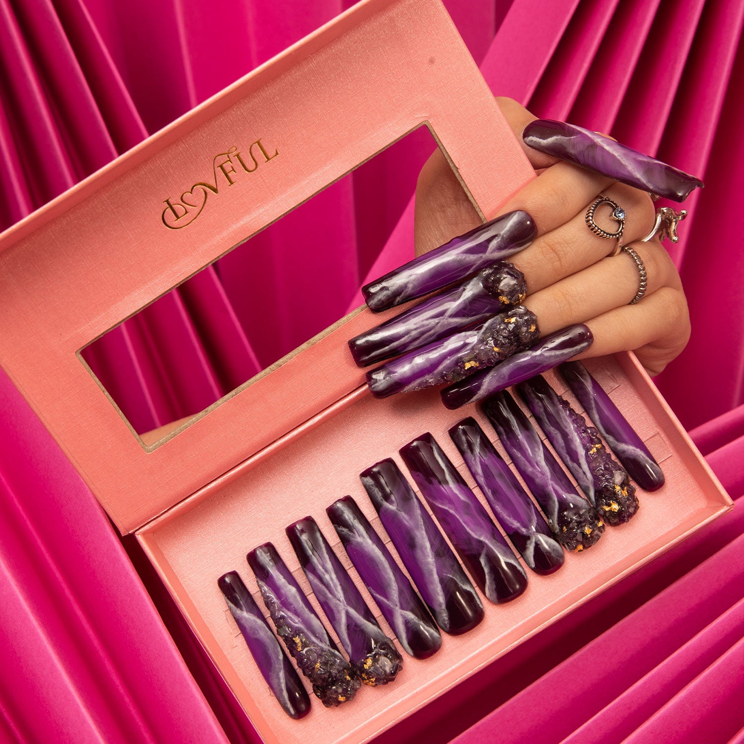 Set of Dream Amethyst press-on nails with elegant purple and black marbled design in a pink box, with a hand wearing matching nails against a draped pink fabric backdrop.