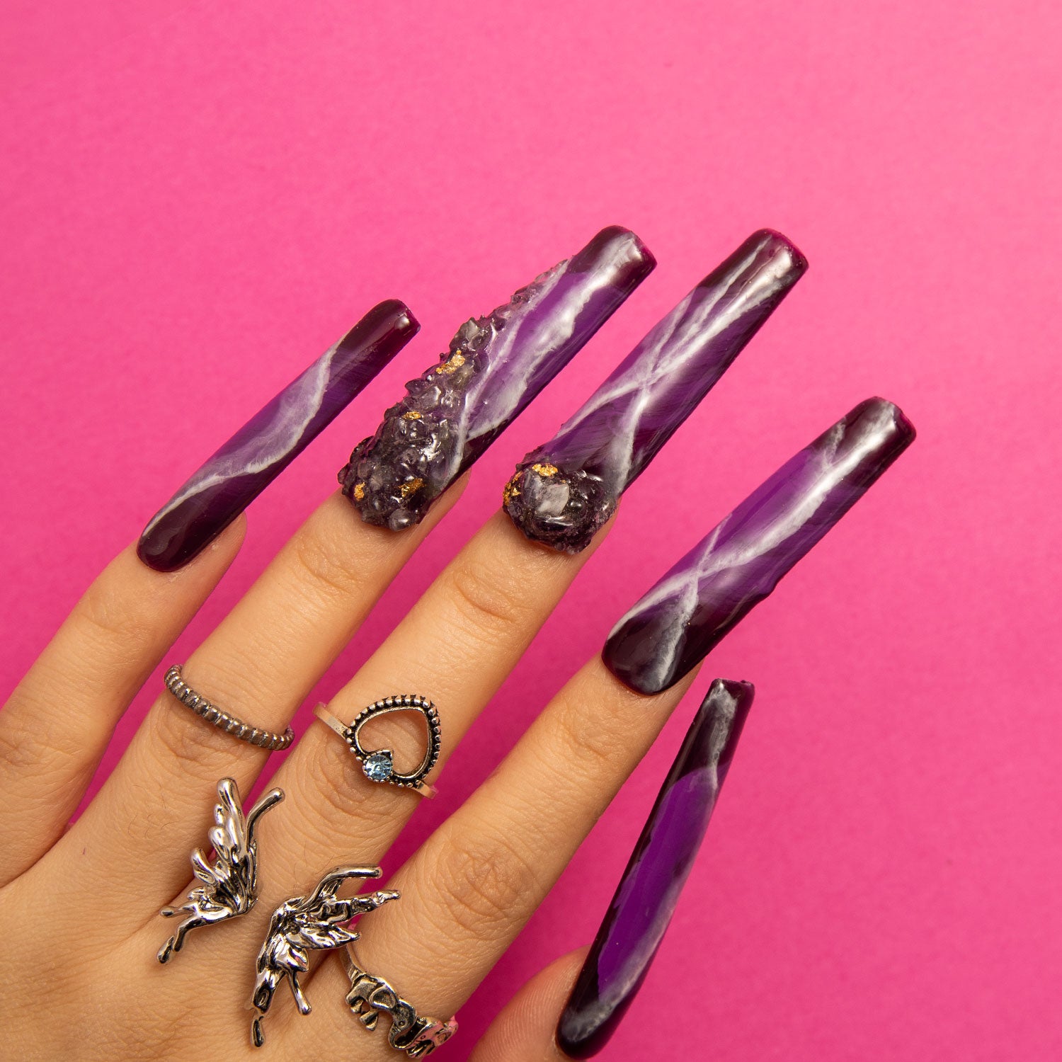 Hand showcasing Dream Amethyst press-on square nails with intricate purple designs and amethyst-like embellishments, accessorized with silver rings, on a pink background.
