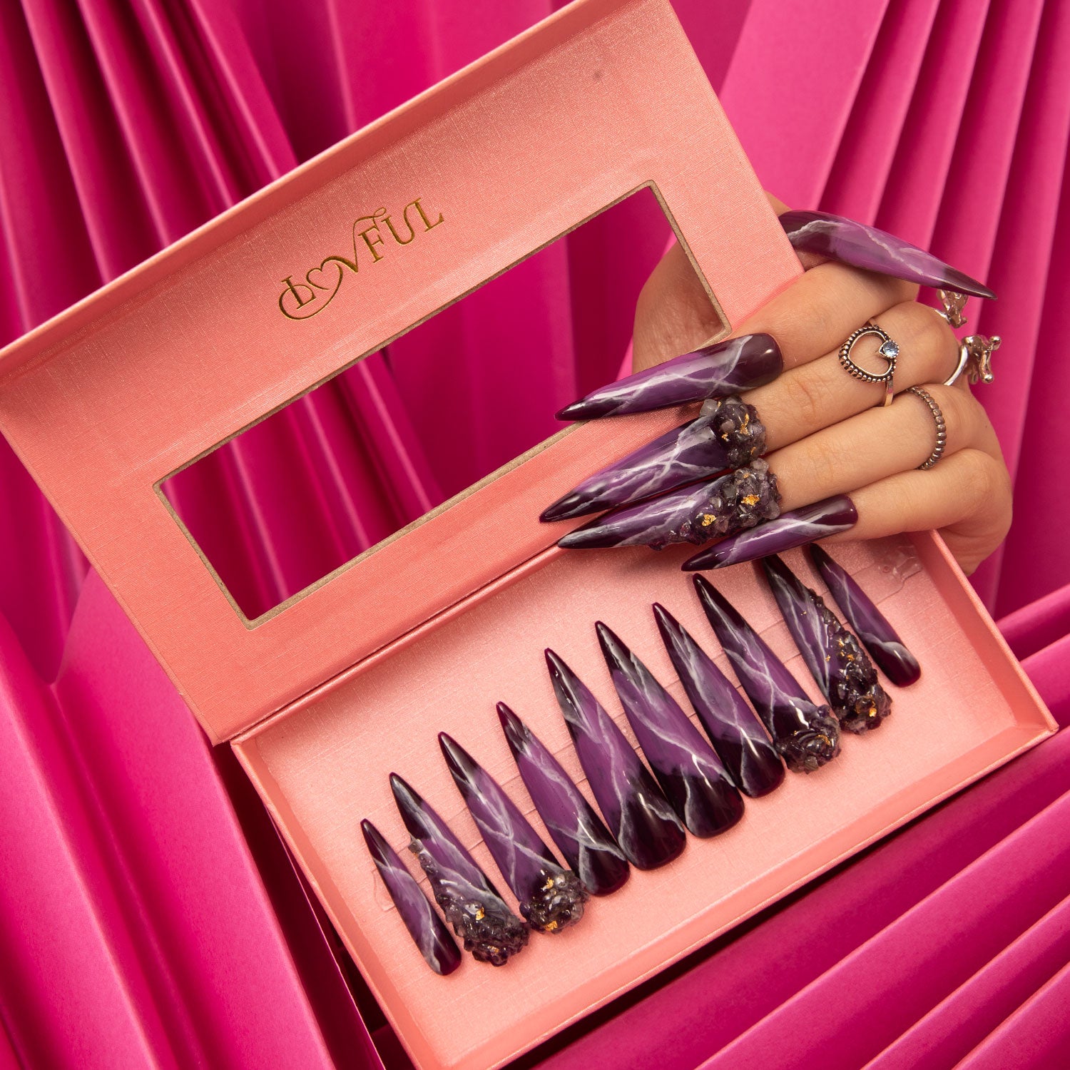 Dream Amethyst Press-On Nails from Lovful in stiletto shape, featuring intricate purple and black designs with celestial artwork, displayed in a pink box. A hand wearing the same nails is adorned with rings, against a vibrant pink fabric background.