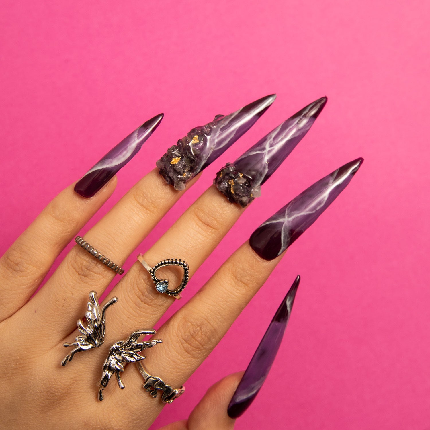 Hand wearing Dream Amethyst stiletto-shaped press-on nails with purple and white designs, adorned with intricate details. The hand also has multiple distinctive rings, set against a bright pink background.